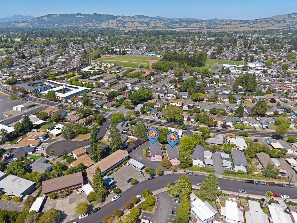 Aerial view of a residential neighborhood with houses, streets, and trees, with a park and sports field in the background, and hills in the distance.