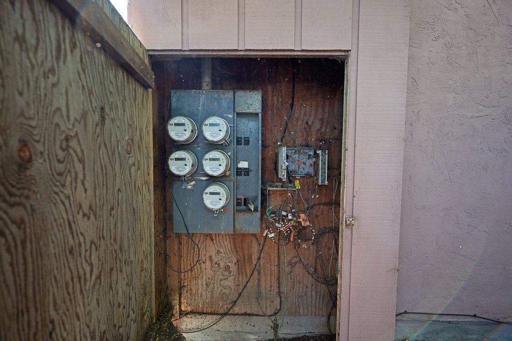 Inside an outdoor electrical meter box with four digital meters, an electrical panel, and wiring on a wooden backboard.