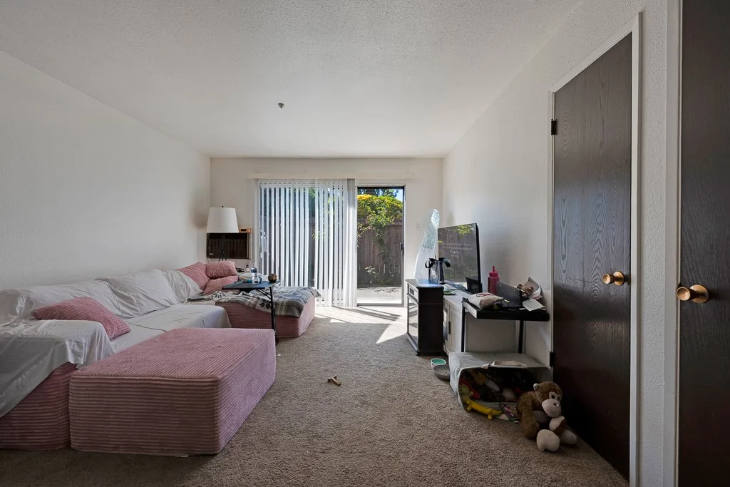 Living room with a white sofa and pink ottoman, television on a black stand, sliding glass door with vertical blinds, and various small items scattered around.