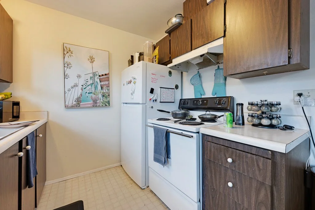 Small kitchen with white refrigerator, wooden cabinets, white countertop, stove with cookware, spice jars, chalkboard, and wall-mounted oven mitts.