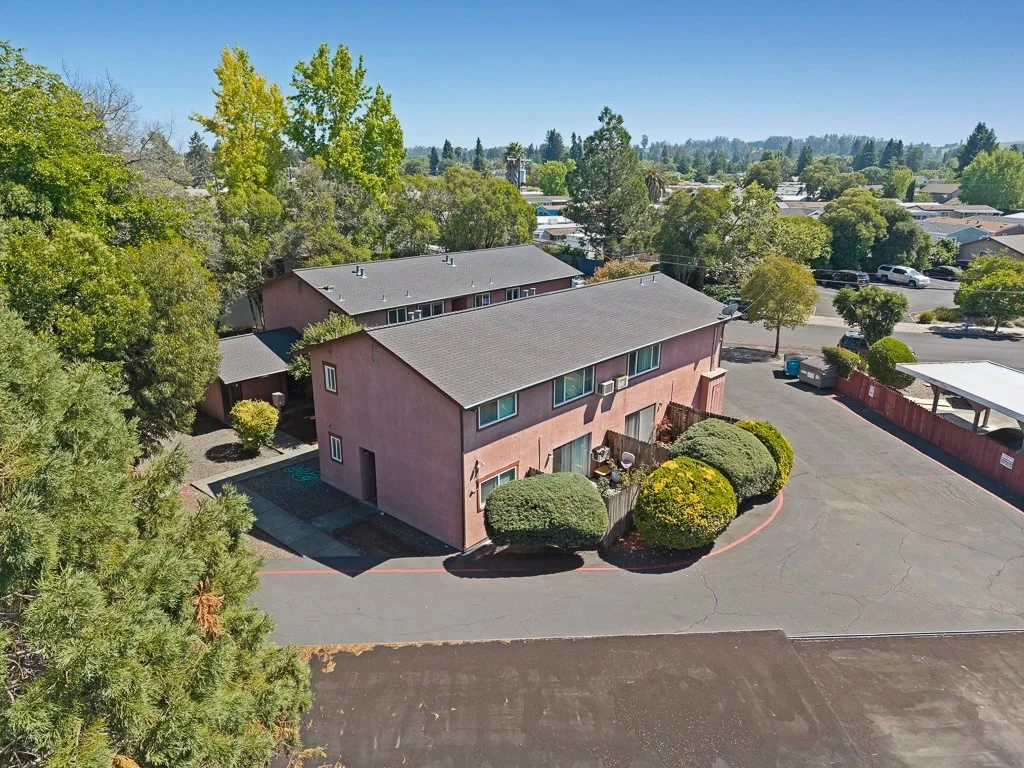 Aerial view of a two-story pink apartment building with bushes and trees, located in a residential neighborhood with parking lots and other buildings.
