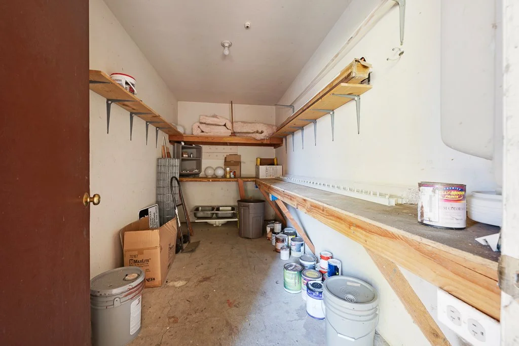 A small storage room with shelves on the walls holding paint cans, boxes, and other supplies. The floor is cluttered with more paint cans and buckets, and there is a trash can and some boxed items in the back corner.