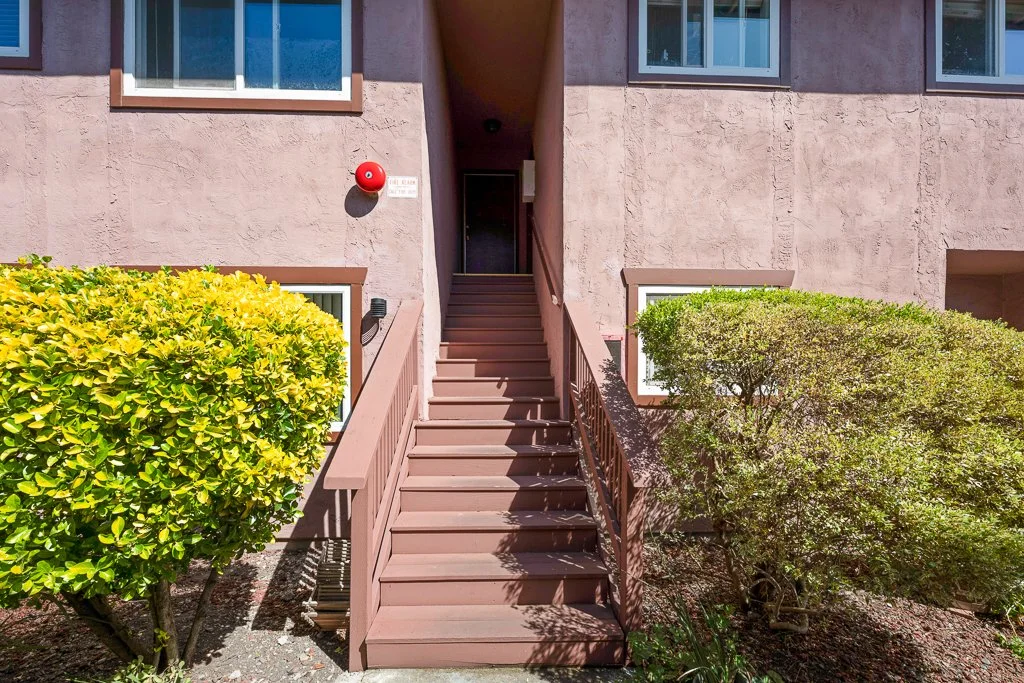 Exterior staircase leading to an upper floor of a pink apartment building, flanked by green bushes and a red alarm bell on the wall.