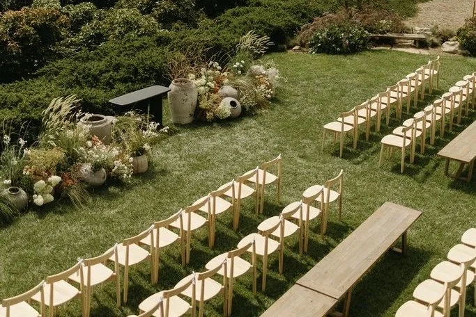 Outdoor garden setting with rows of wooden chairs arranged facing a floral aisle, decorated with large vases and plants.