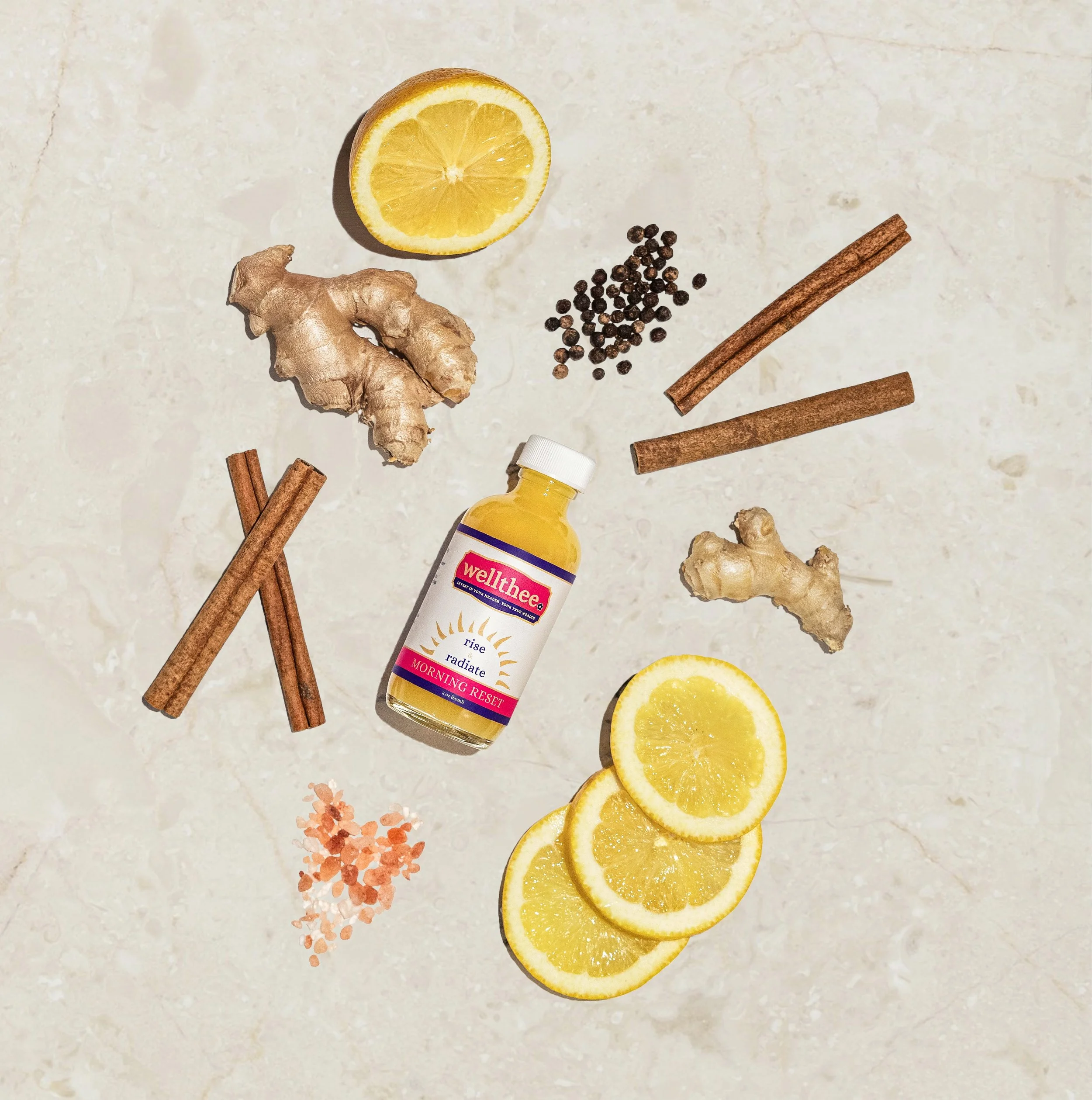 Half lemon, ginger root, black peppercorns, cinnamon sticks, pink Himalayan salt, wellthee nootropic supplement bottle, sliced lemon, salt flakes, and a yellow supplement bottle on a light-colored surface.