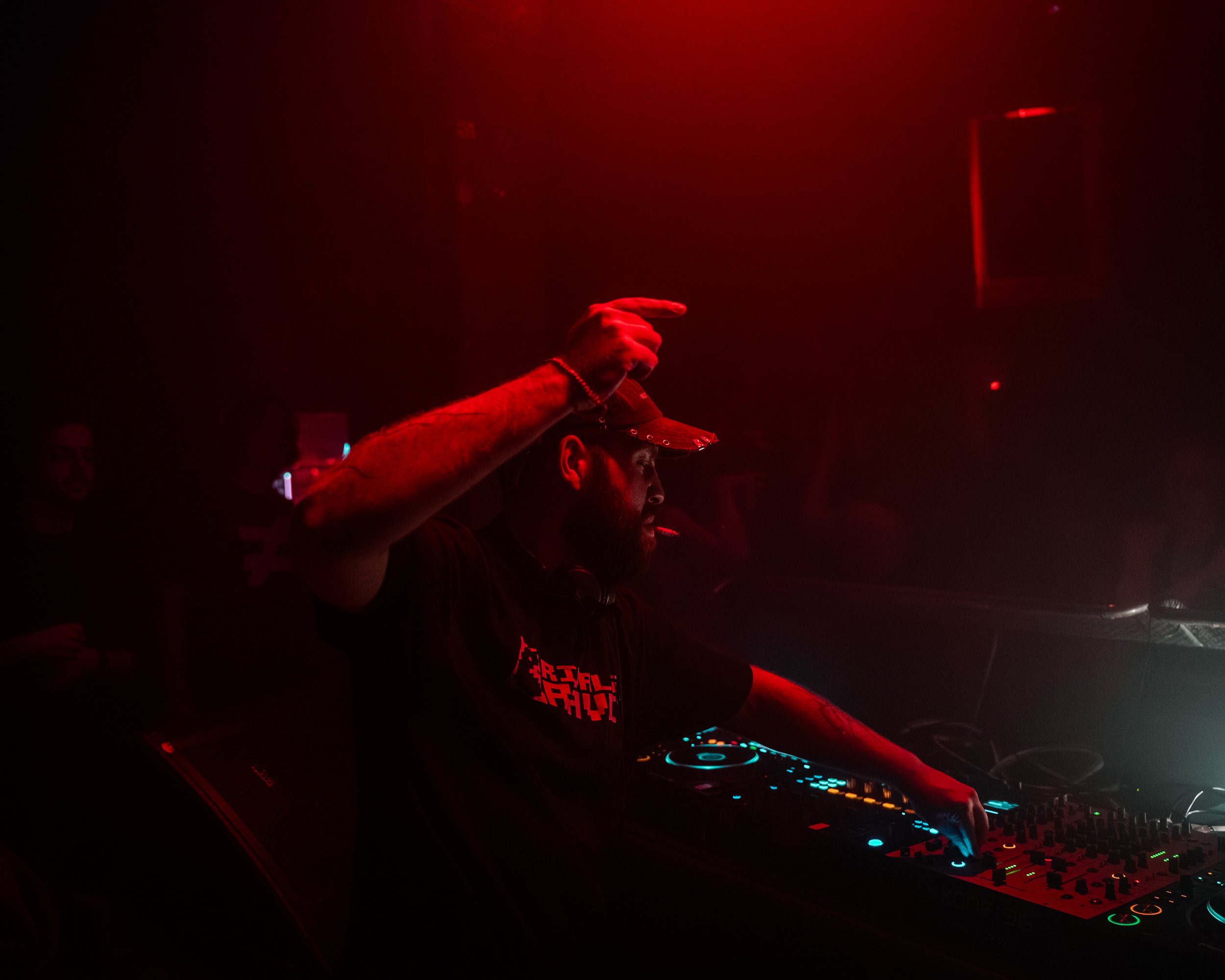 Ein DJ mit Headphones und DJ-Equipment in einer dunklen, rot beleuchteten Umgebung, vermutlich bei einem Club oder einer Party.