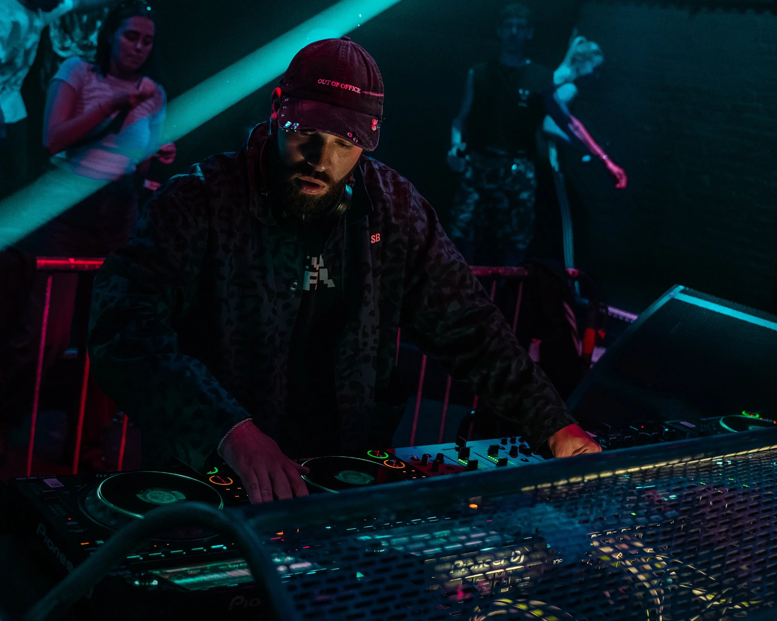 DJ spielt auf einem beleuchteten DJ-Pult in einer Bar oder einem Club, umgeben von mehreren Menschen, bei dunkler Atmosphäre mit farbigem Licht.