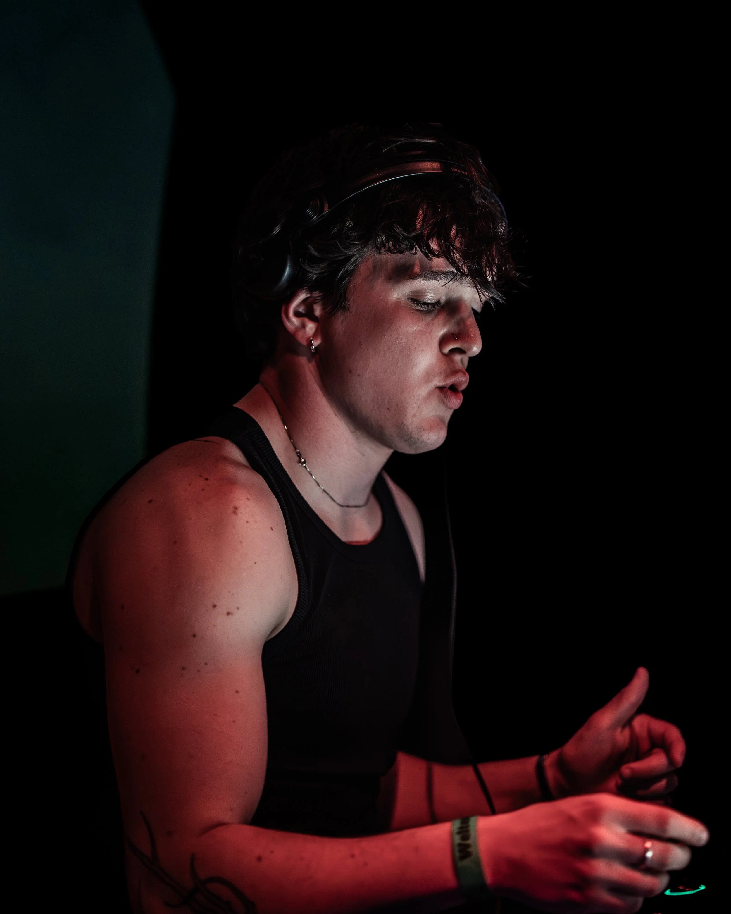 Nahaufnahme einer Person mit dunklen, lockigen Haaren, die in einem dunklen Raum mit rotem Licht arbeitet, vermutlich beim DJing oder Musikproduktion, mit Kopfhörern und am Gerät.