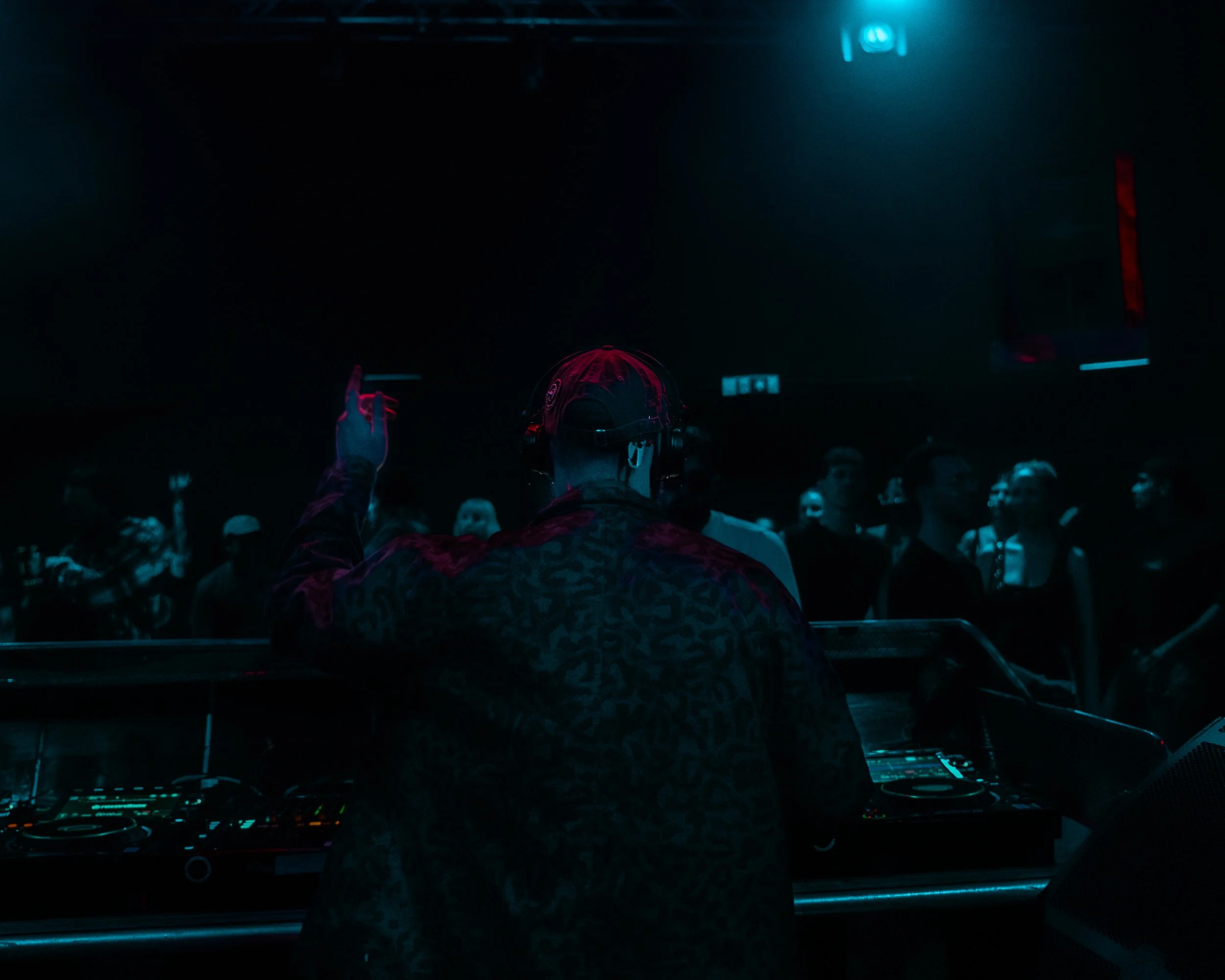 Blick auf eine dunkle Menschenmenge bei einer Veranstaltung, im Vordergrund ein DJ mit Kopfhörern und einer DJ-Konsole, im Hintergrund blaue Lichtstrahlen.