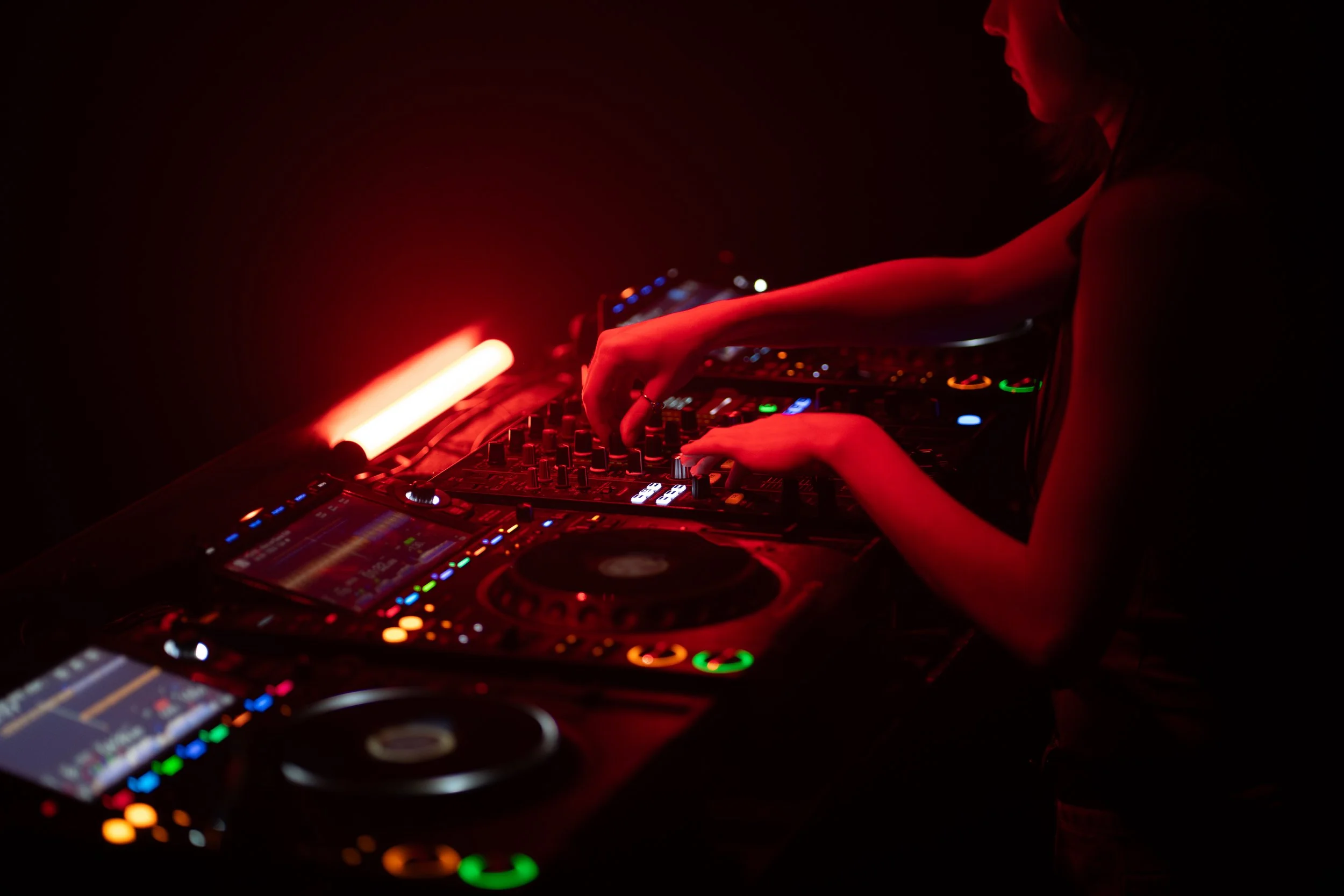 Eine Person mit dunkler Kleidung, die in einer dunklen Umgebung auf einem DJ-Pult mit bunten Lichtern arbeitet.
