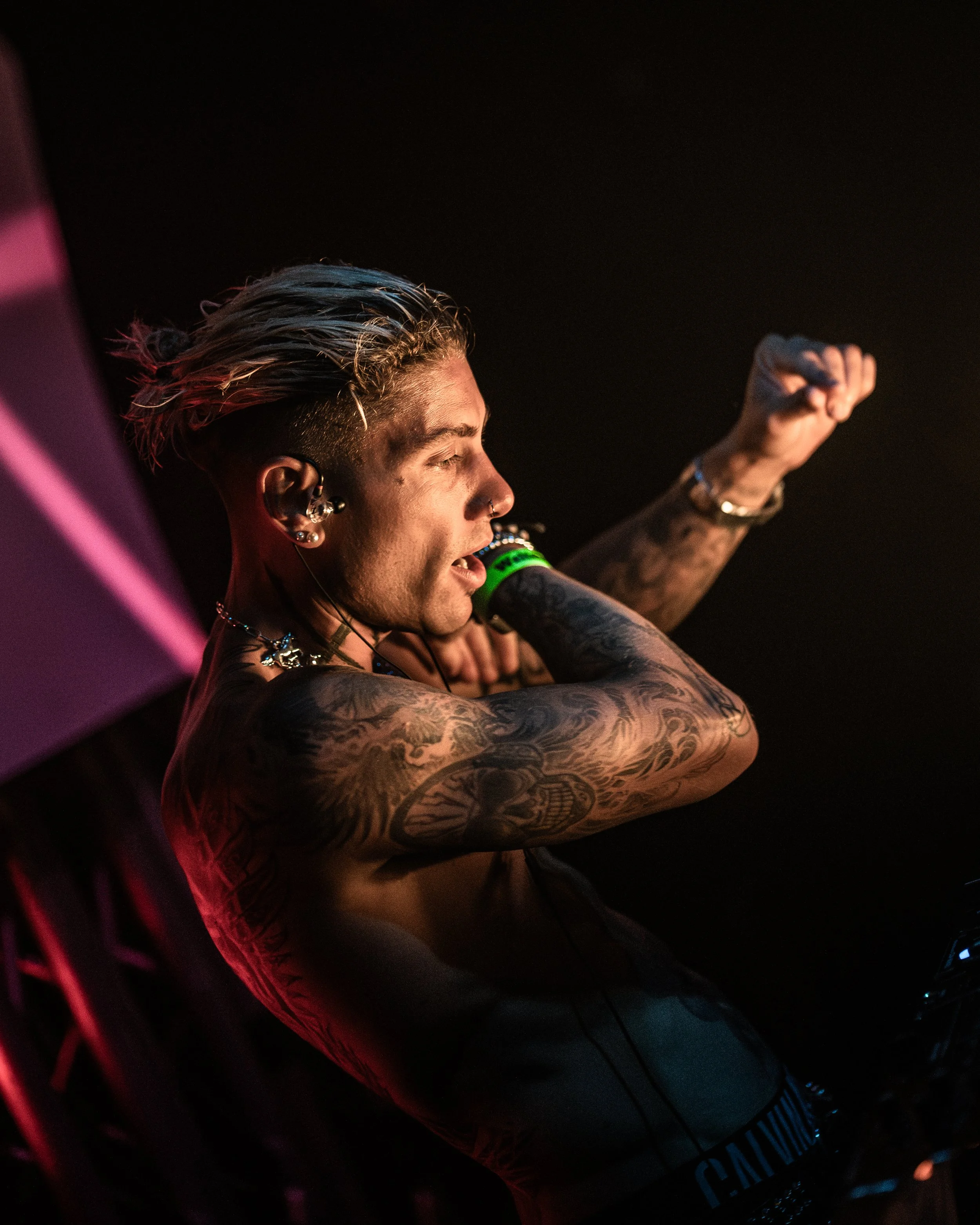 Seitenansicht eines jungen männlichen Musikers mit Tattoos, Piercings, dunklen Haaren mit blonden Spitzen und einem Ohrring, der in einem dunklen Raum mit schwarzem Hintergrund singt oder spricht in ein Mikrofon.