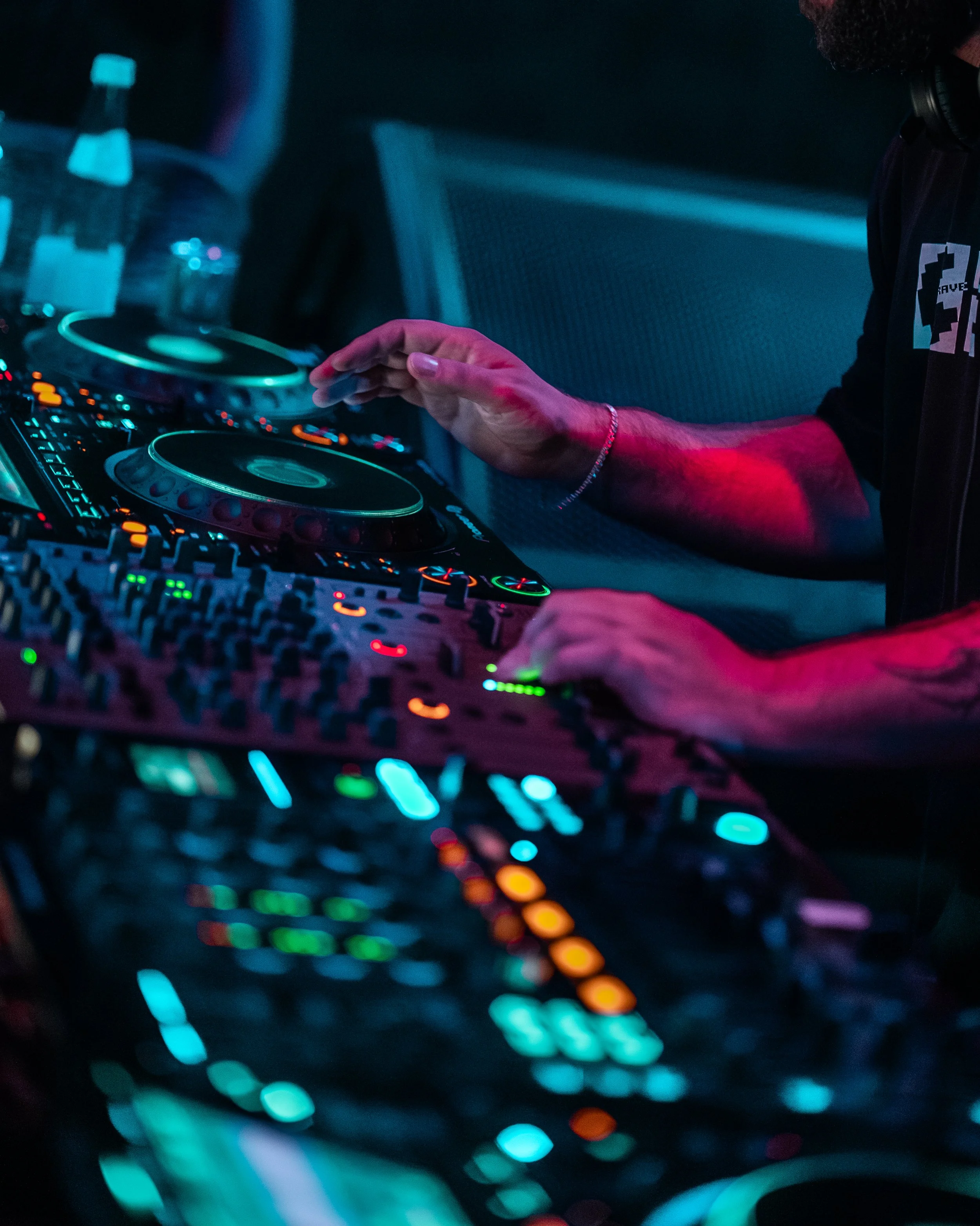 Nahaufnahme eines DJs beim Auflegen an einem DJ-Controller in dunklem, mit bunten Lichtern erleuchteten Raum.