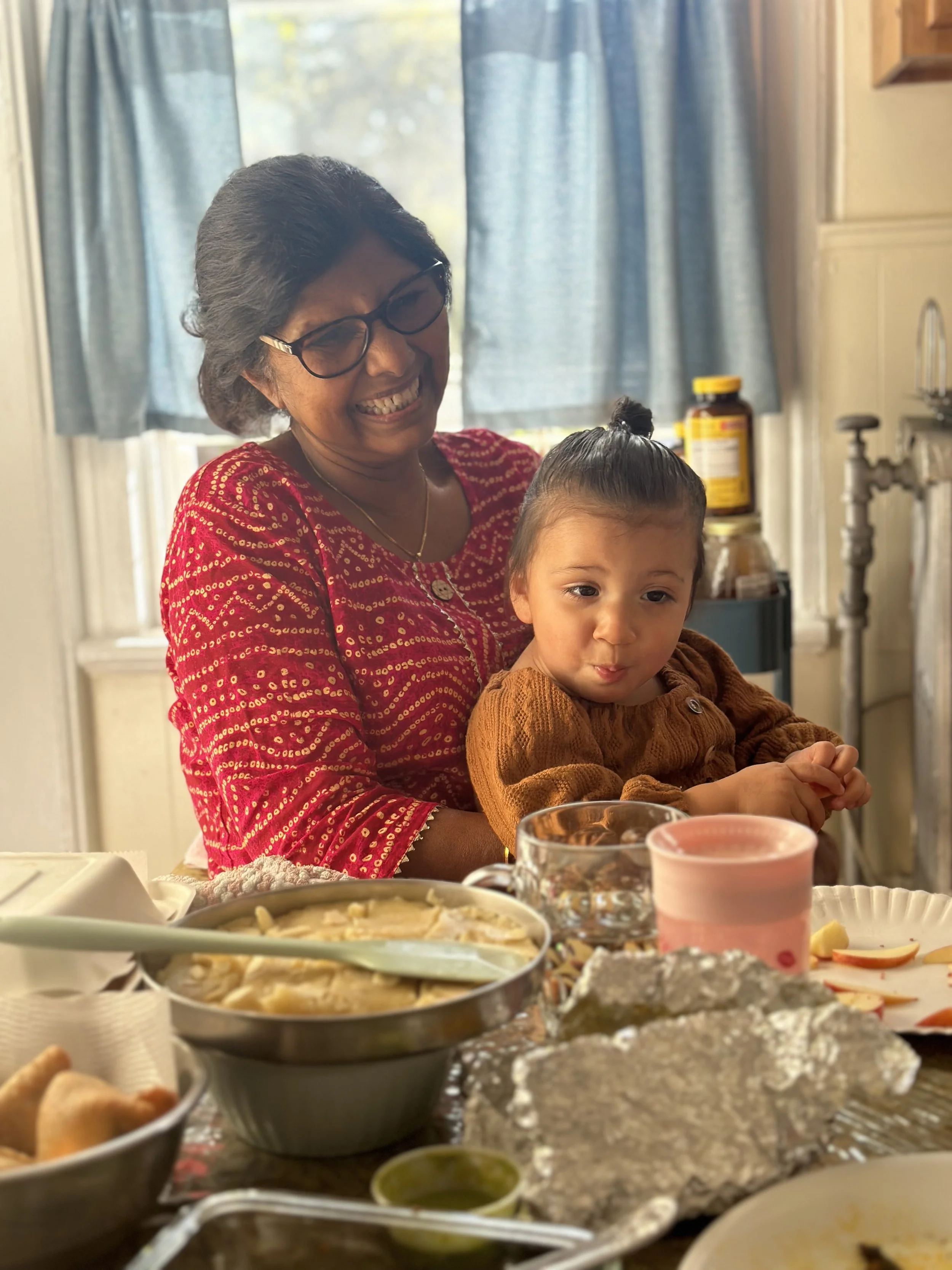 Woman and young girl at a table during a meal, with various dishes and food items in the foreground, in a kitchen setting with blue curtains in the background.