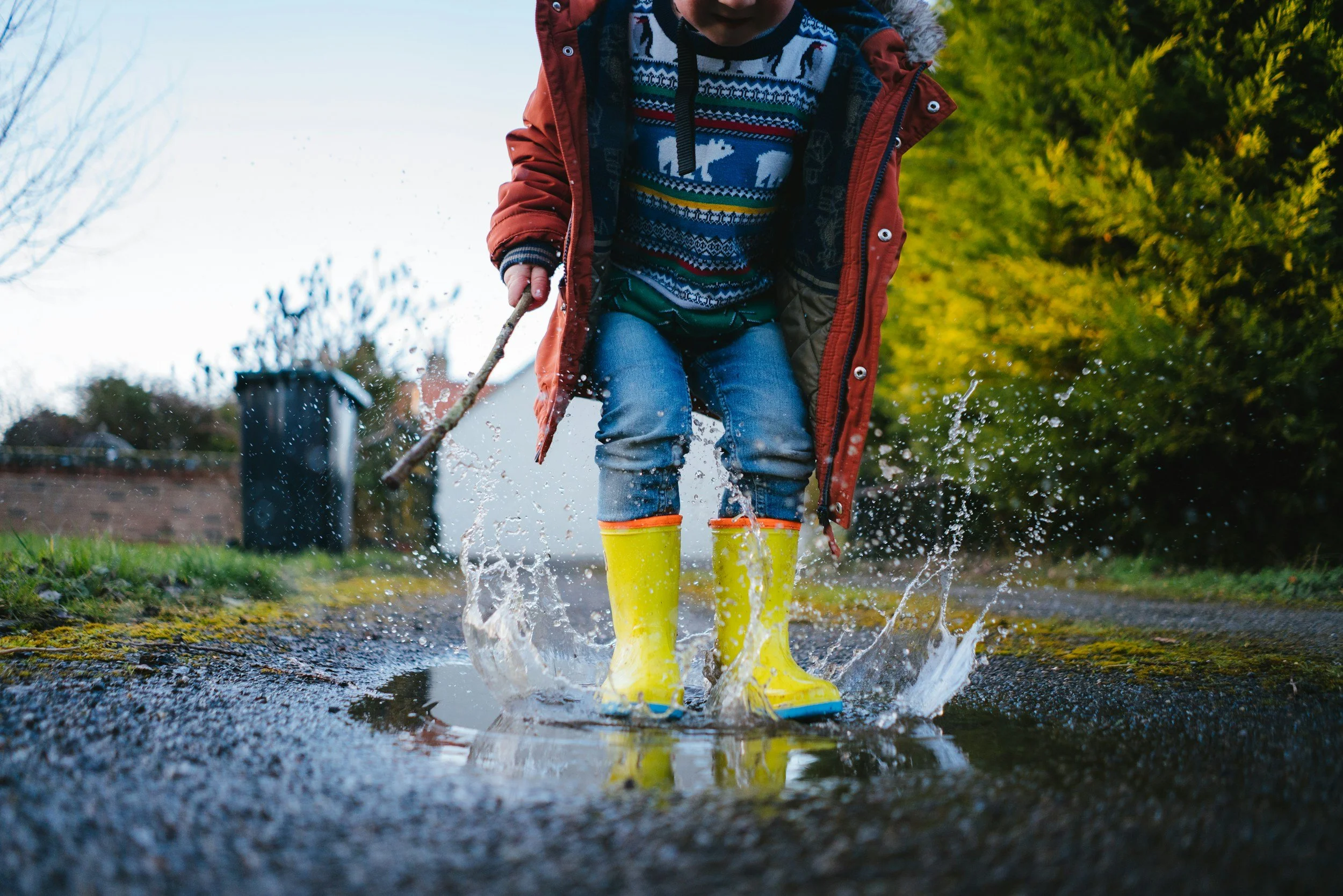Child outdoors wearing yellow rain boots, walking through a puddle, splashing water, dressed in colorful sweater, red coat, and blue jeans, with greenery and houses in the background.