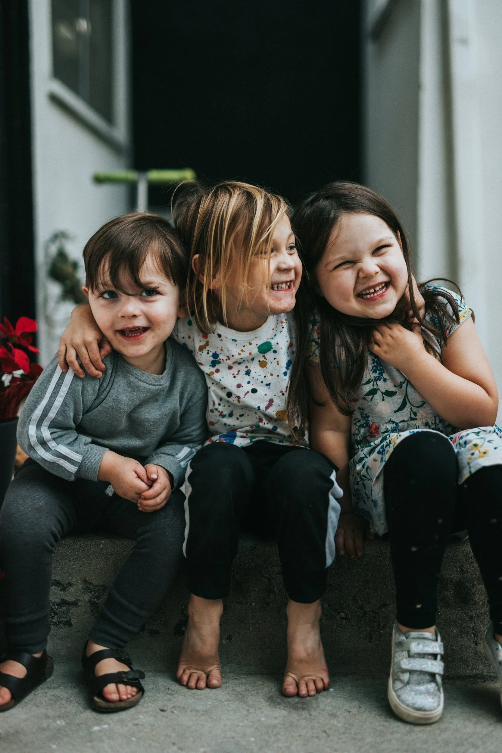Three children sitting closely together, smiling and laughing. A girl with dark hair and a girl with light brown hair in the middle, and a boy with dark hair on the left. They are outside, near a building with an overhang, and some plants are visible in the background.