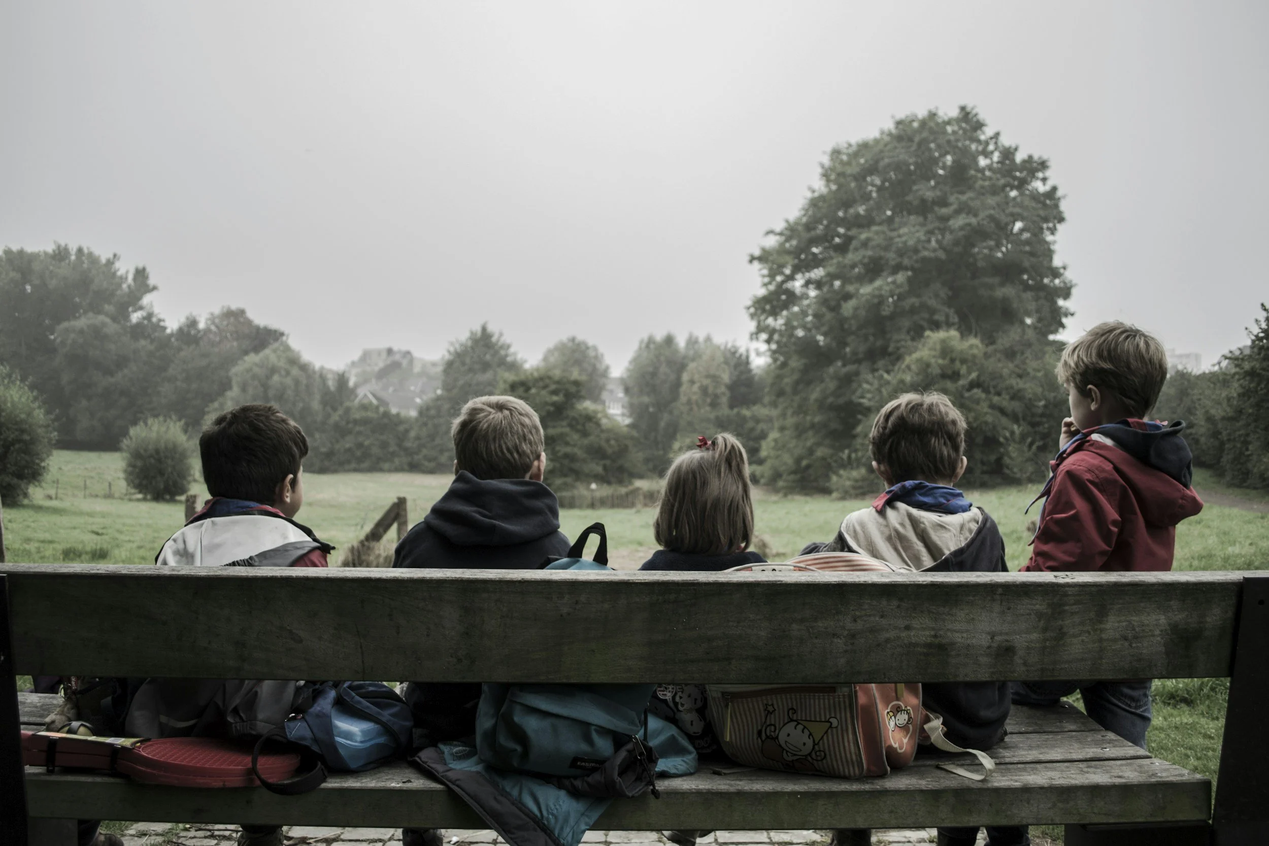 Children sitting on a bench outdoors, facing a green field with trees and cloudy sky.
