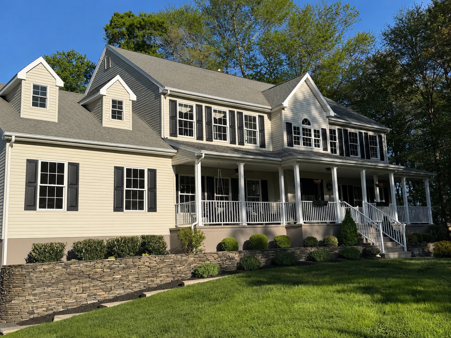A large two-story house with beige siding, black shutters, and a gray roof, featuring a covered front porch with white railings and steps leading down to a well-maintained lawn, surrounded by green bushes and tall trees in the background.