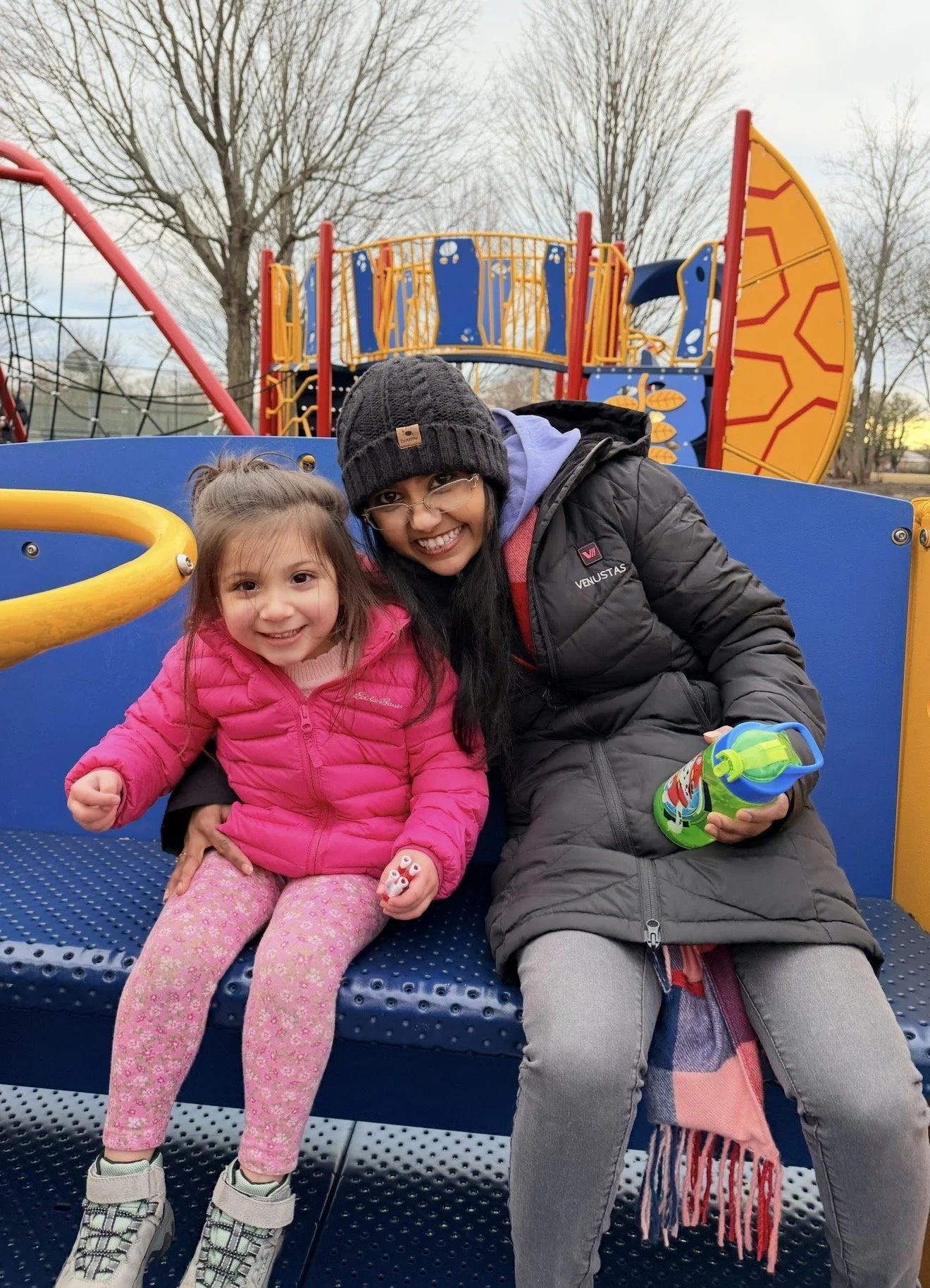 A woman and a young girl sitting on a blue bench at a playground, smiling at the camera. The girl is wearing a pink puffy jacket and pink patterned pants, holding a small toy. The woman is dressed in a black coat, gray pants, and a knitted beanie, holding a colorful water bottle. Playground equipment and leafless trees are visible in the background.