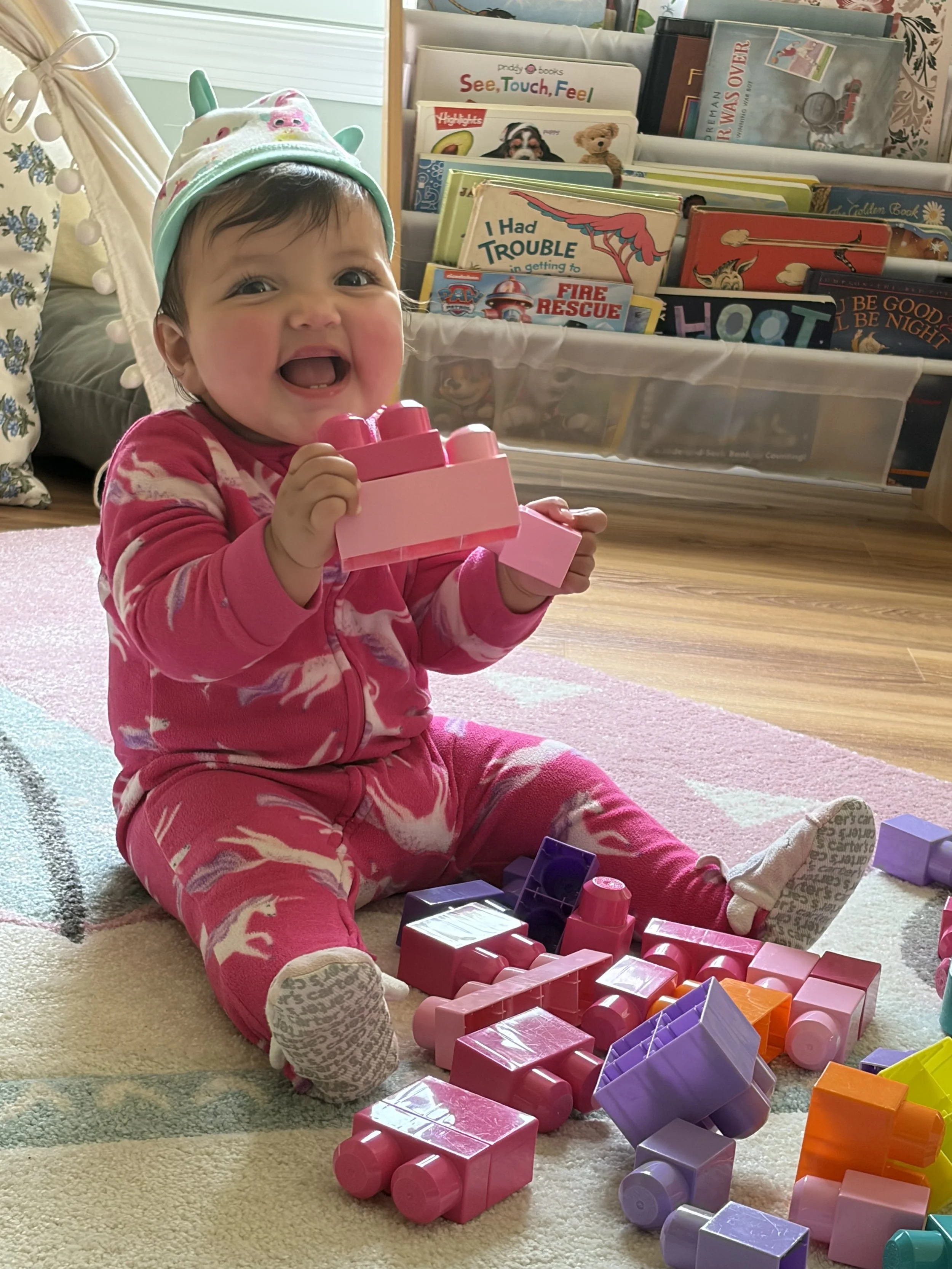 A smiling toddler girl sitting on a rug, holding a pink toy truck, surrounded by colorful toy building blocks, with a bookshelf filled with children's books in the background.