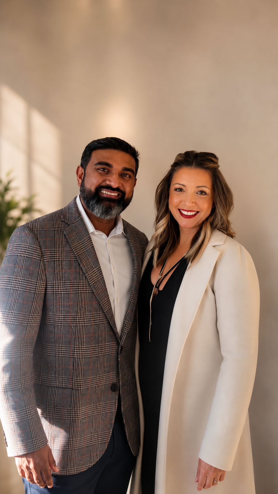 A man and woman standing side by side, smiling, dressed in business attire, with a neutral background and natural light.