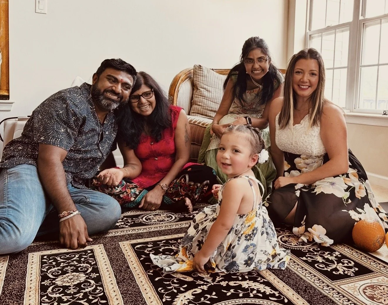 Group of six diverse people, including children, gathered in a living room, sitting on a patterned rug around a small pumpkin. They are smiling and posing for a photo near a window with sunlight coming in.