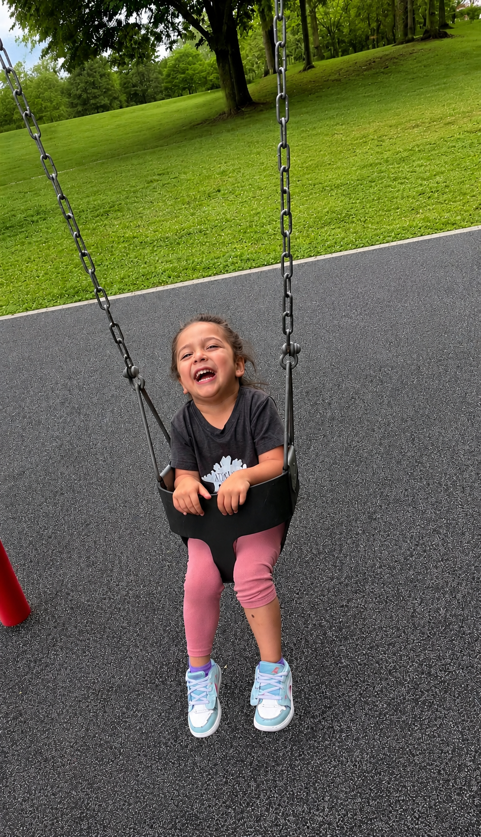 A young girl with brown hair smiling and sitting in a black bucket swing at a playground, wearing a black t-shirt, pink leggings, and white sneakers. Green grass, trees, and an overcast sky are in the background.