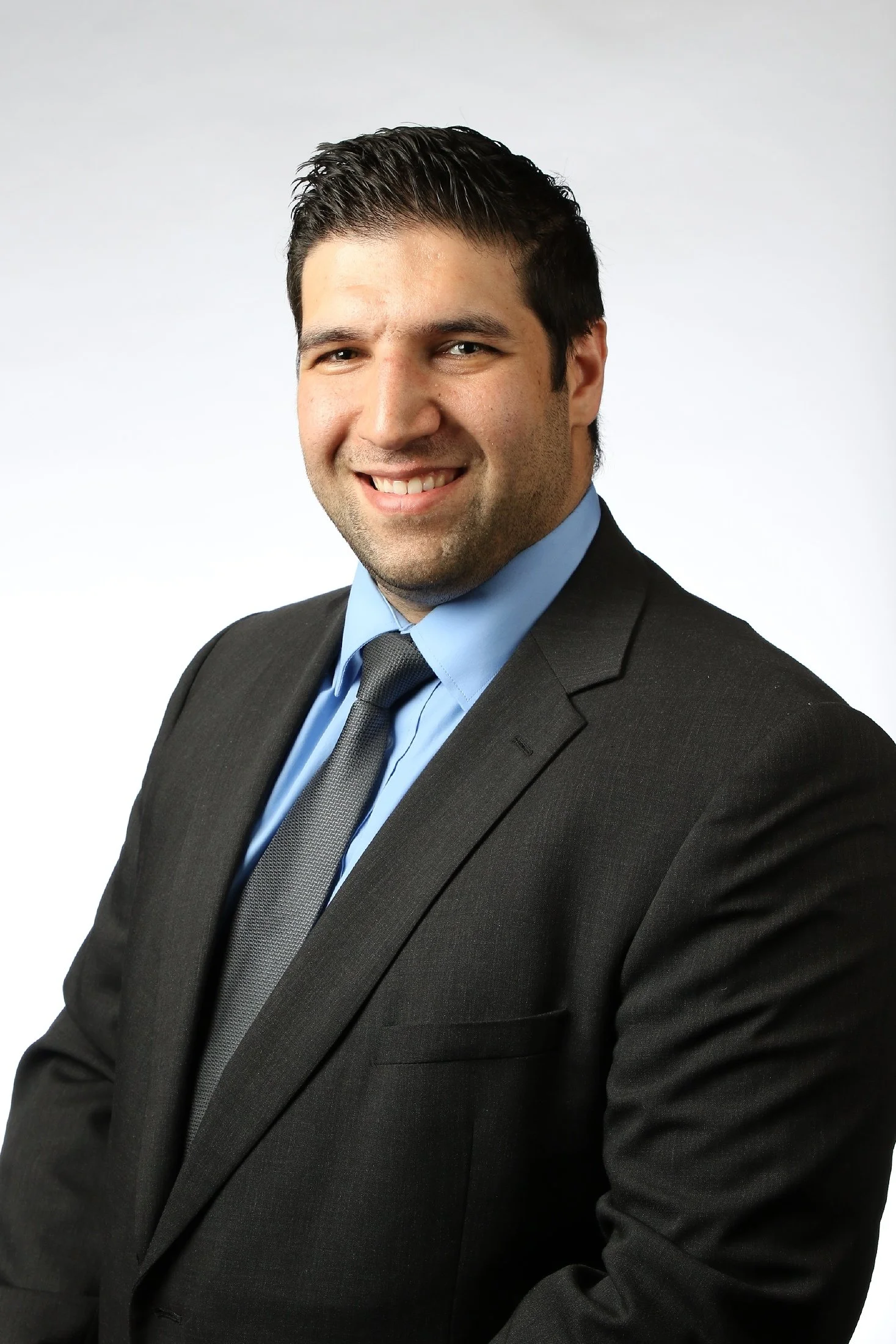 A close-up professional portrait of a smiling man wearing a dark suit, light blue shirt, and gray tie against a plain light gray background.