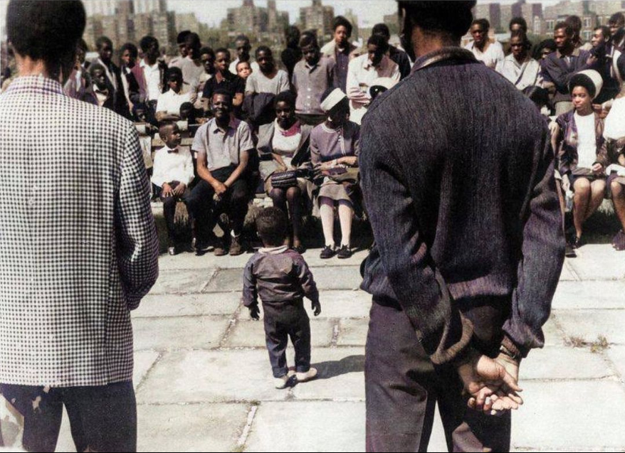 Group of people gathered outdoors, listening to two men speaking; a small child stands between them facing the audience.