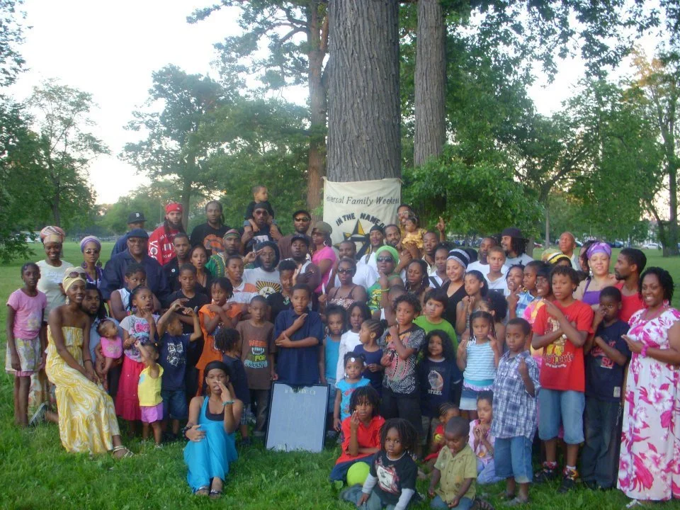 A large group of children and adults gathered outdoors near trees for a community event, with a banner in the background that mentions 'Family Weekend'.