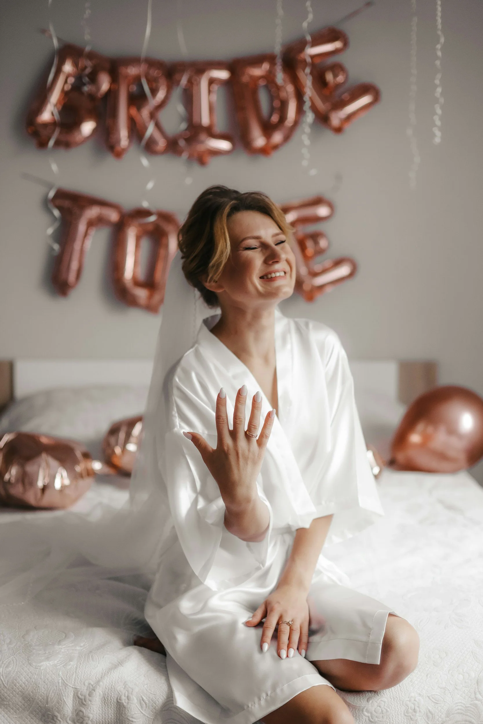 A woman in a white satin robe sitting on a bed with balloons in the background that spell out "LOVE" and "WIFE" in rose gold. She is smiling with her eyes closed and showing her wedding ring.