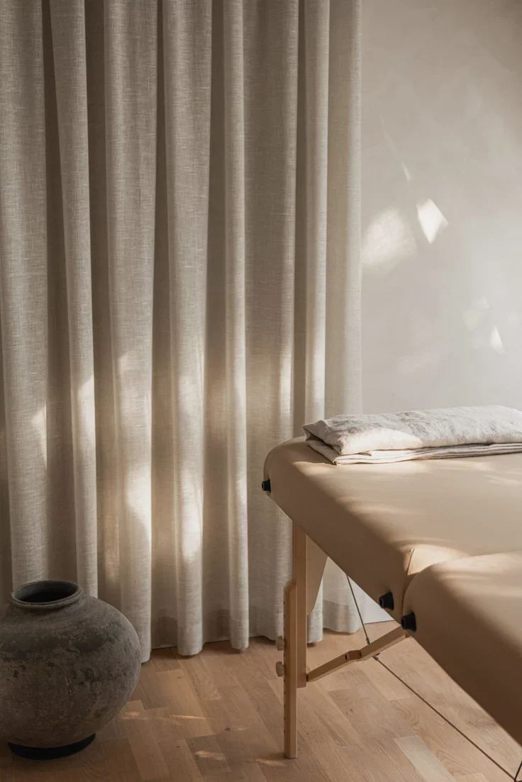 A massage table with folded towels, a large ceramic vase, beige curtains, and a wooden floor in a room.