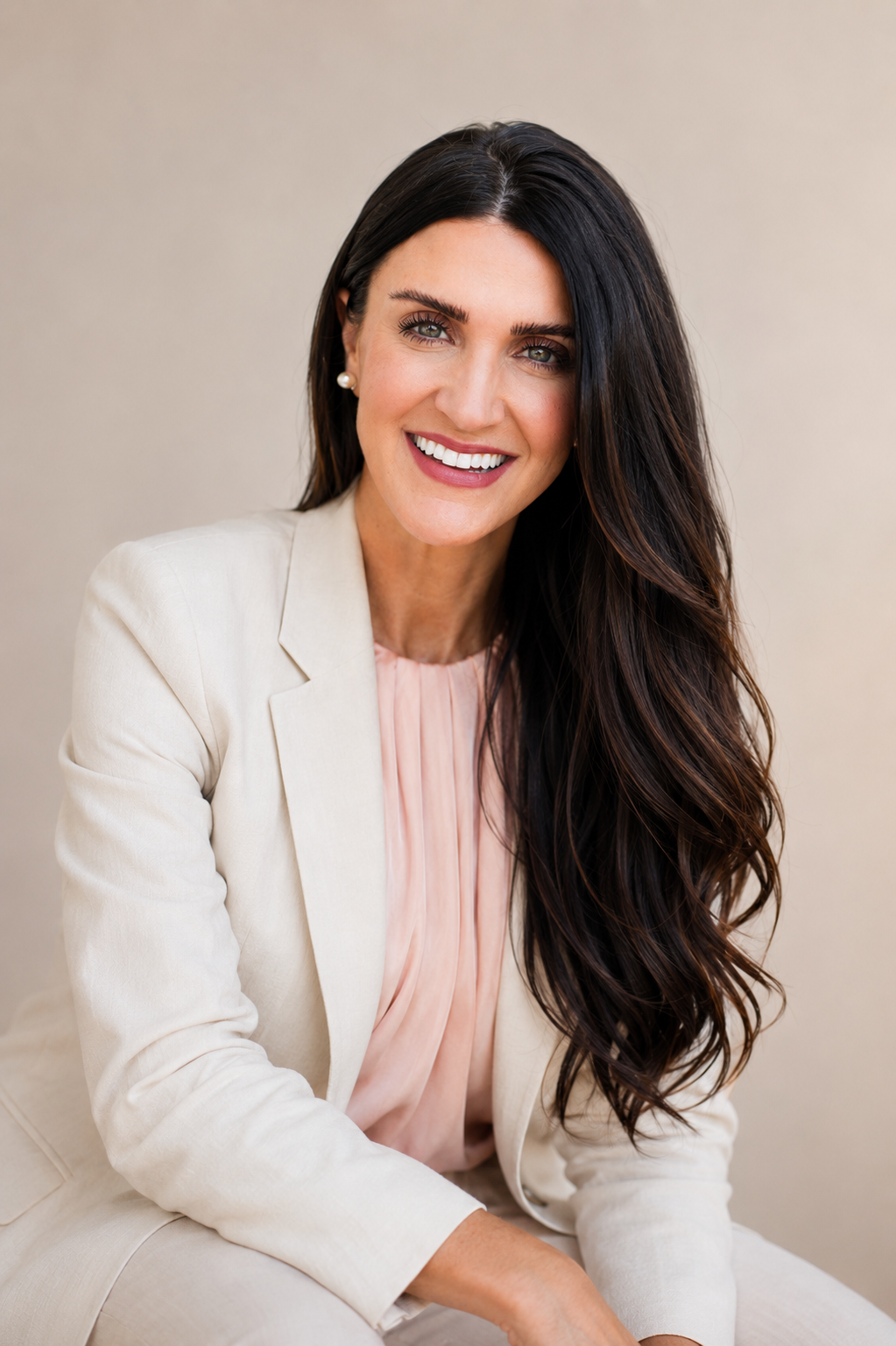 A woman with long dark brown hair and blue eyes smiling, wearing a cream-colored blazer, a light pink blouse, and pearl earrings, sitting against a plain beige background.