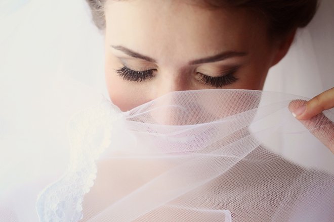 Close-up of a woman with makeup and long eyelashes holding a sheer, white fabric near her face.