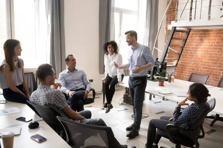 Six people in a modern office space having a discussion or meeting, some standing and some sitting, with a staircase and brick wall in the background.