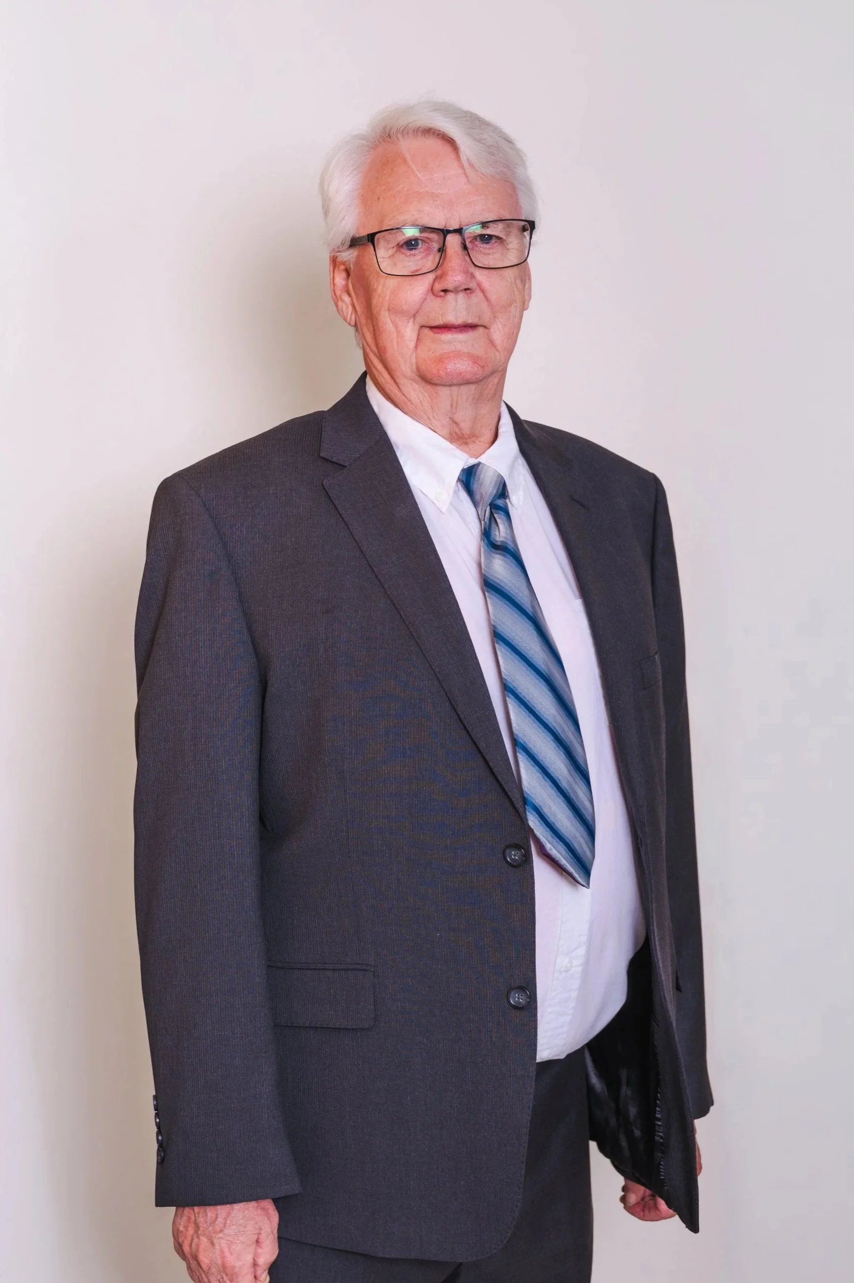 An elderly man with white hair, glasses, and a serious expression, wearing a dark gray suit, white shirt, and striped tie, standing against a plain white background.