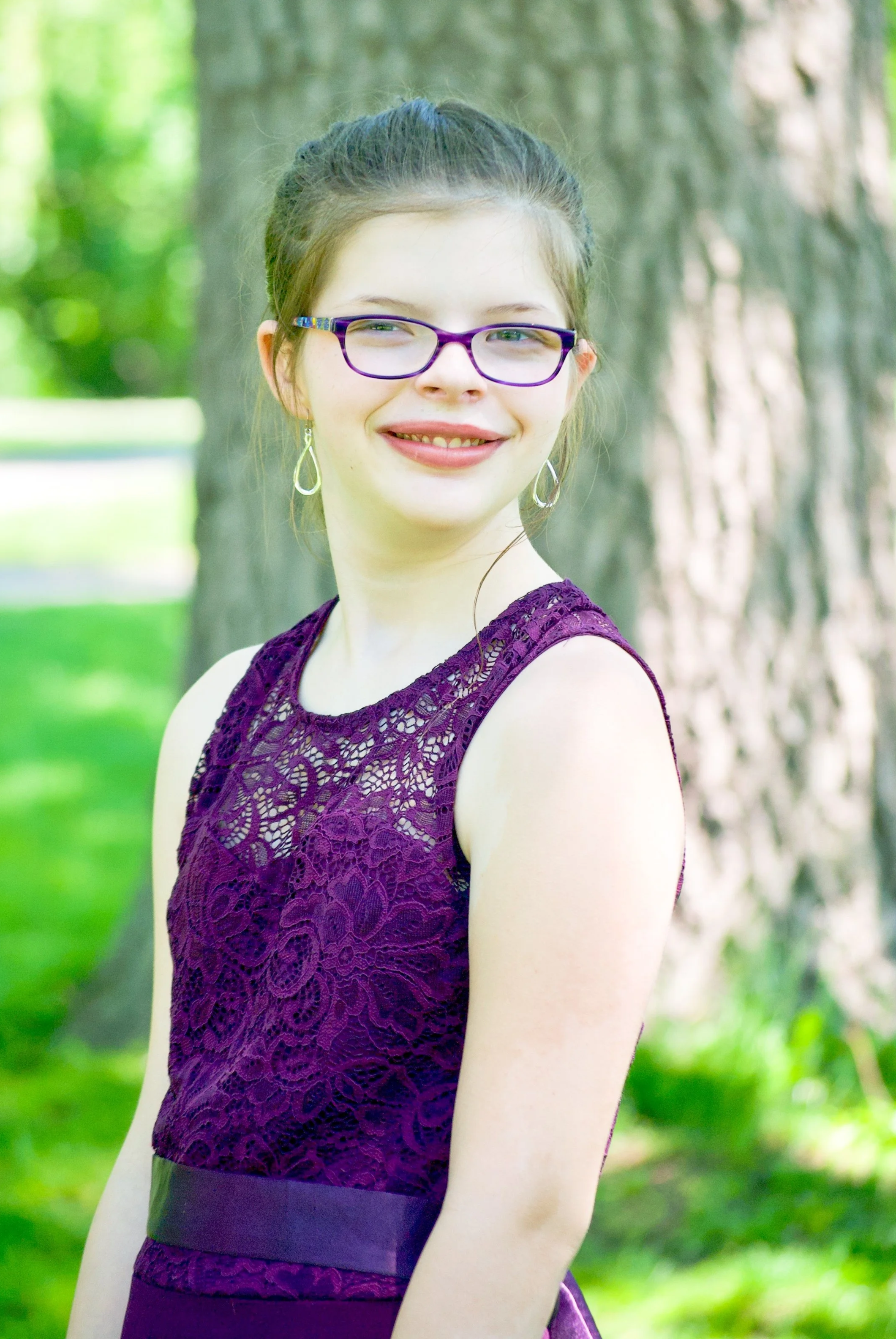 A young woman with glasses, earrings, and a lace purple dress standing outdoors in a park-like setting with trees and greenery in the background.
