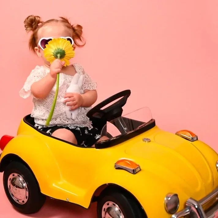 A young girl with curly hair in buns, wearing sunglasses, sitting in a yellow toy car, holding a yellow flower close to her face against a pink background.