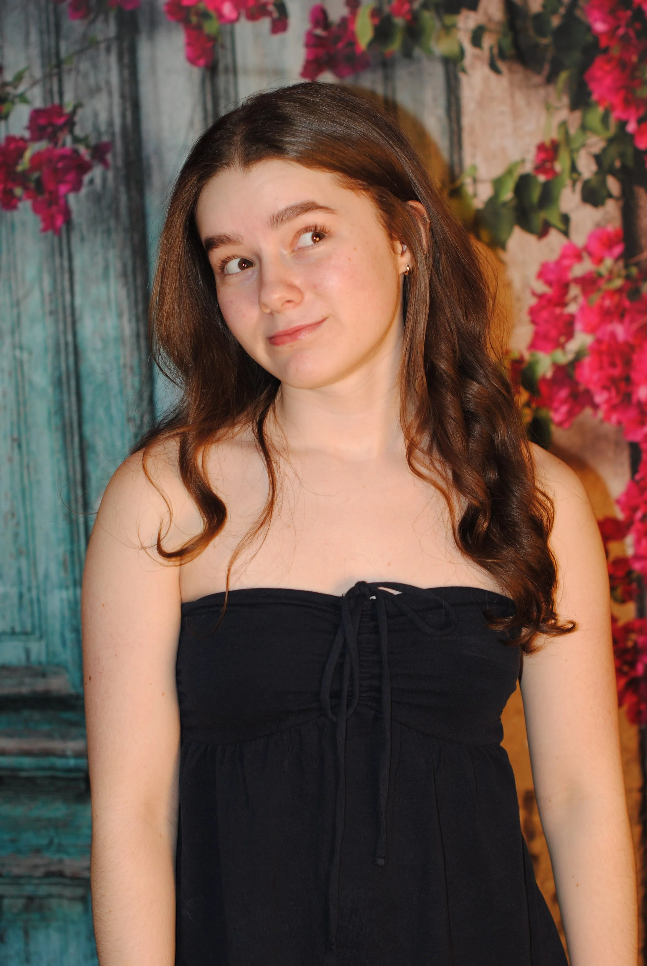 A young woman with brown hair styled in loose curls, wearing a black strapless dress, standing in front of a colorful background of pink bougainvillea flowers and a weathered wooden fence.