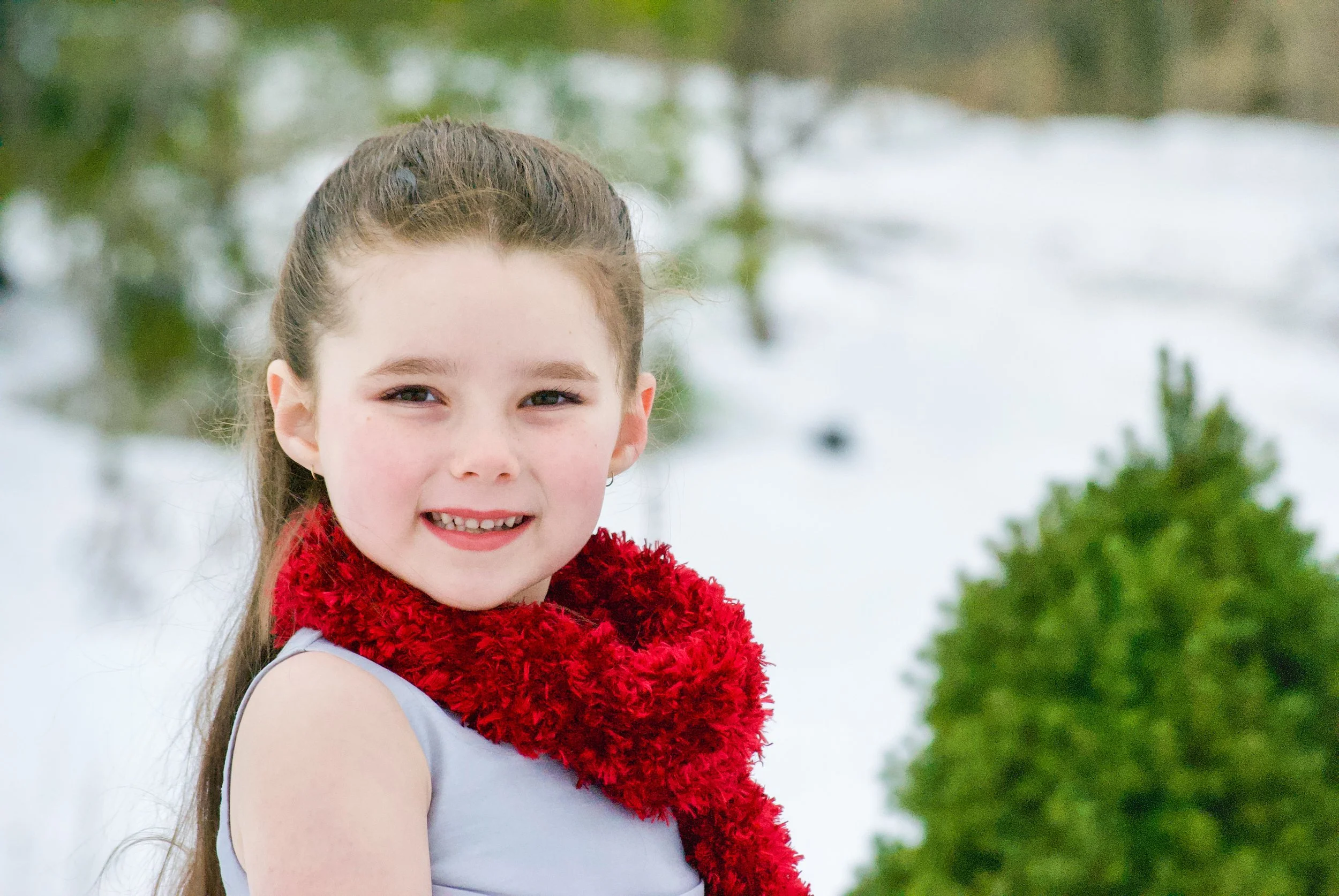 A young girl with long brown hair and a big smile standing outdoors in a snowy landscape with green trees in the background, wearing a white top and a red fuzzy scarf.