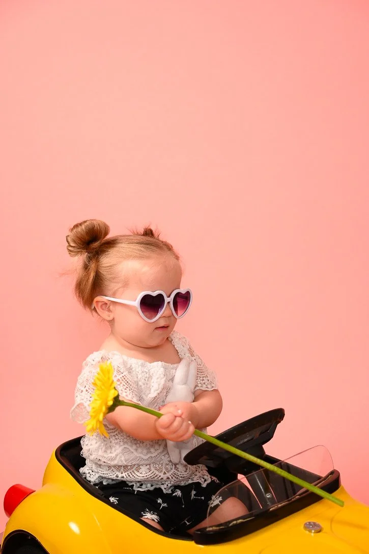 A young girl with heart-shaped sunglasses, wearing a lace top, sitting in a yellow toy car, holding a yellow flower against a pink background.
