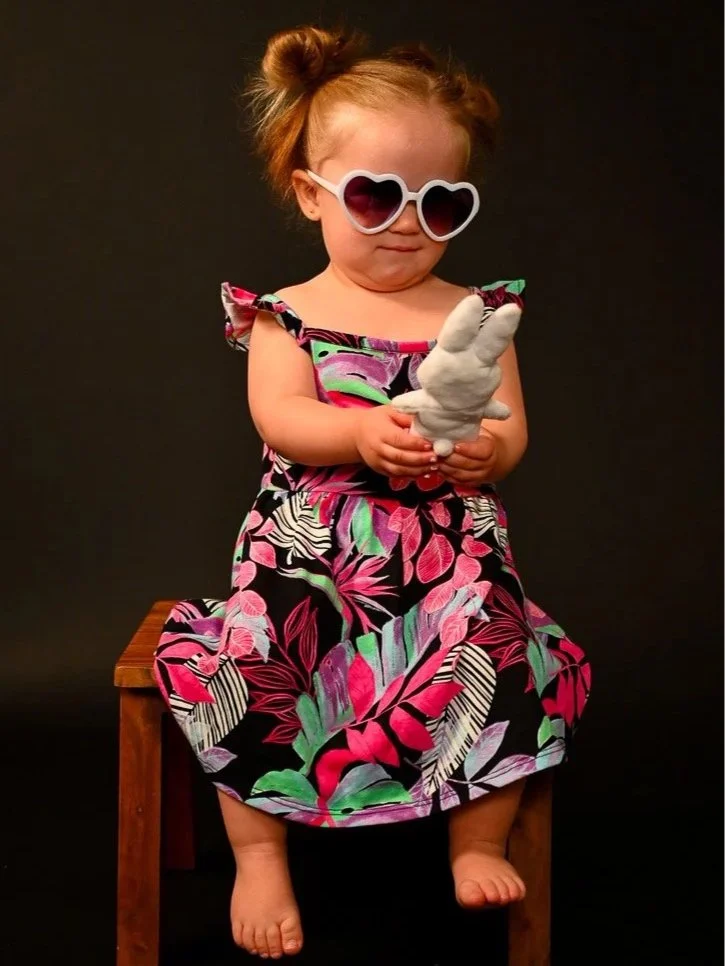 A young girl with red hair styled in two buns, wearing large white sunglasses with heart-shaped lenses, a colorful floral dress, and holding a white stuffed animal, sitting on a small wooden bench against a black background.
