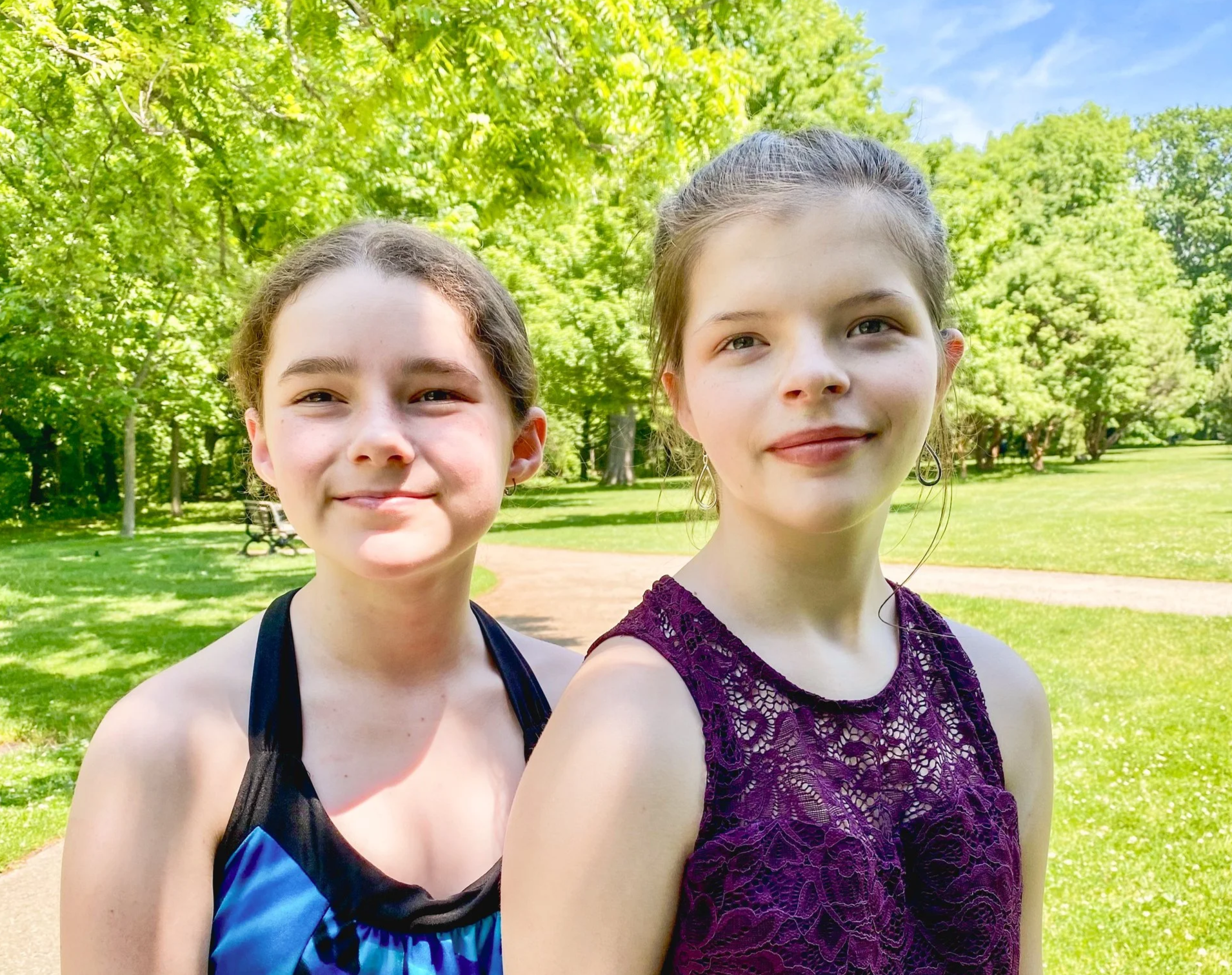 Two young women standing outdoors in a park with lush green trees, smiling at the camera on a sunny day.