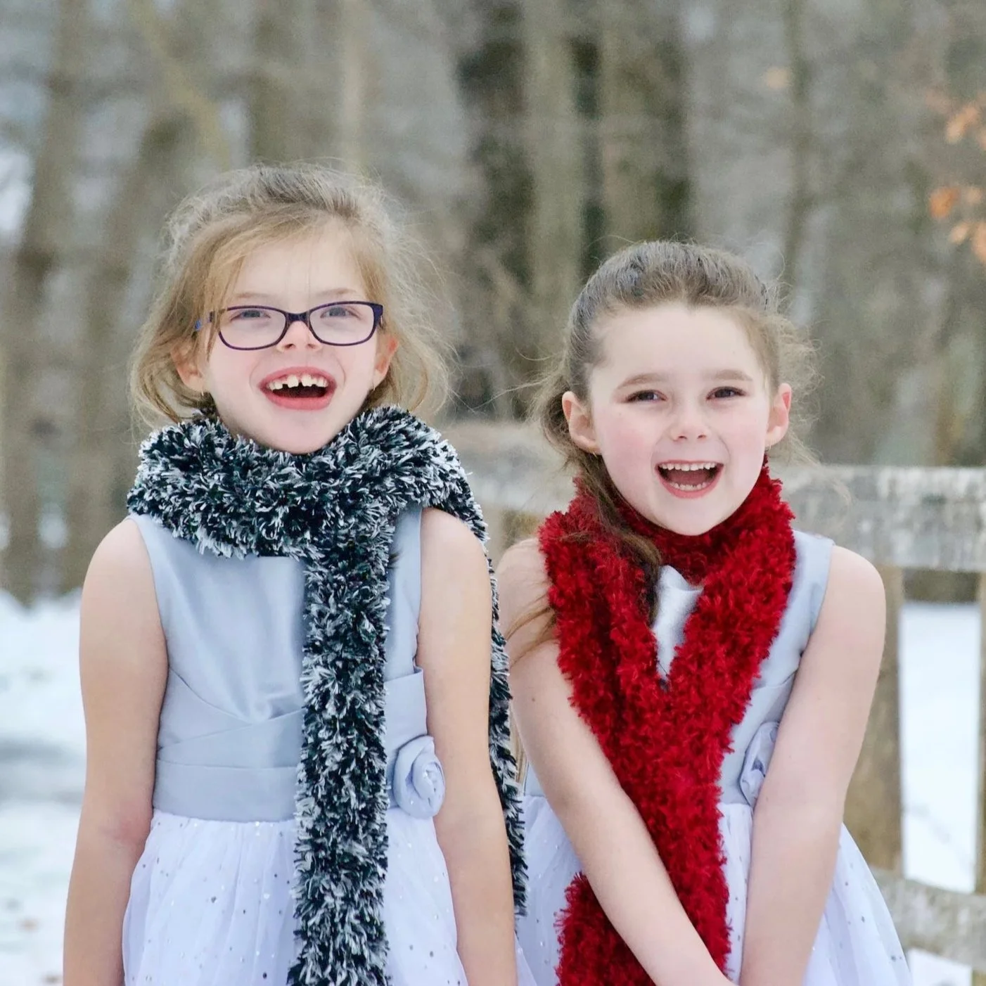 Two young girls wearing white sleeveless dresses and fur scarves, smiling outdoors in a winter setting.