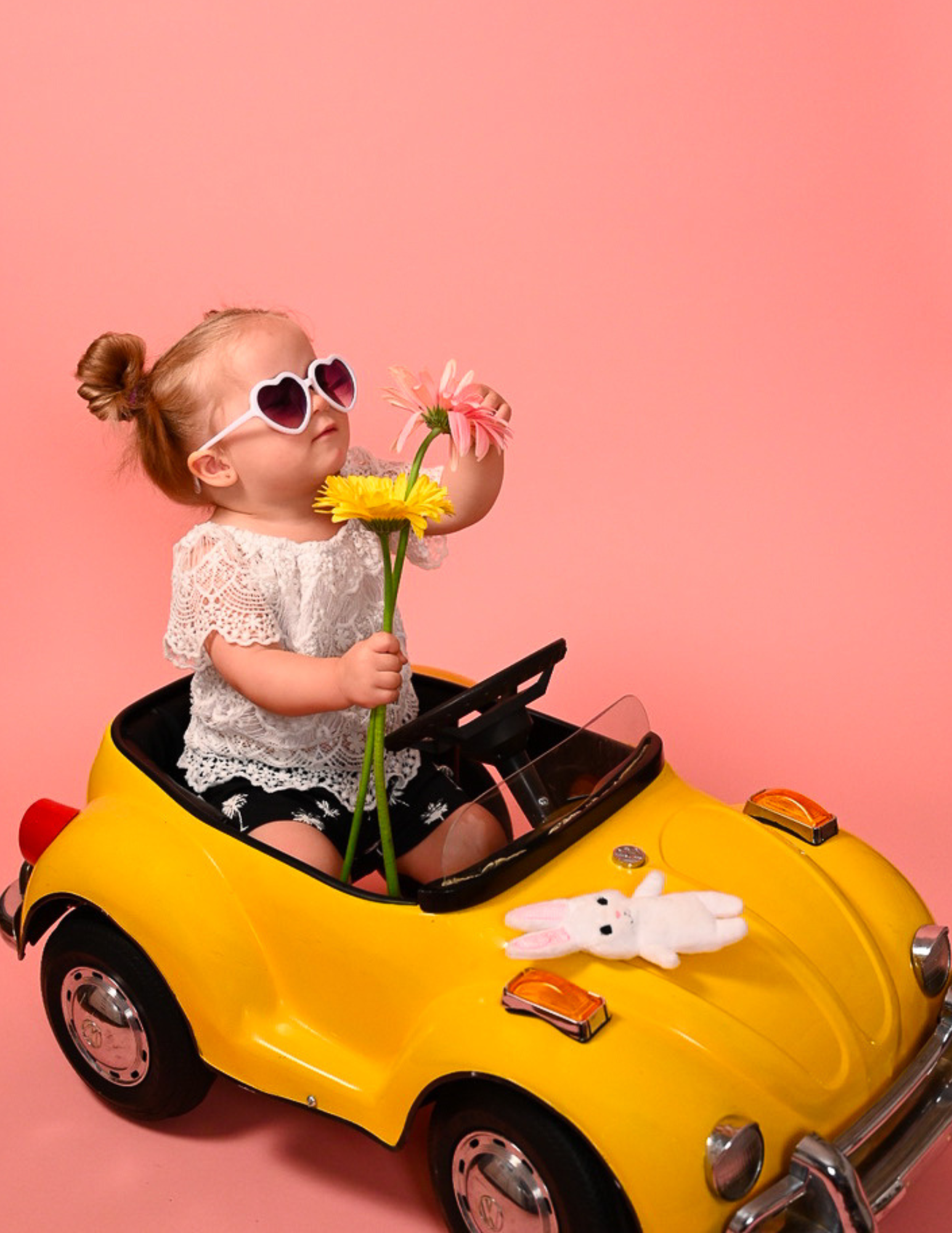 A young girl with red hair in a small yellow toy car, wearing heart-shaped sunglasses, holding a pink flower, with a white bunny stuffed animal on the car's hood, and a pink background.