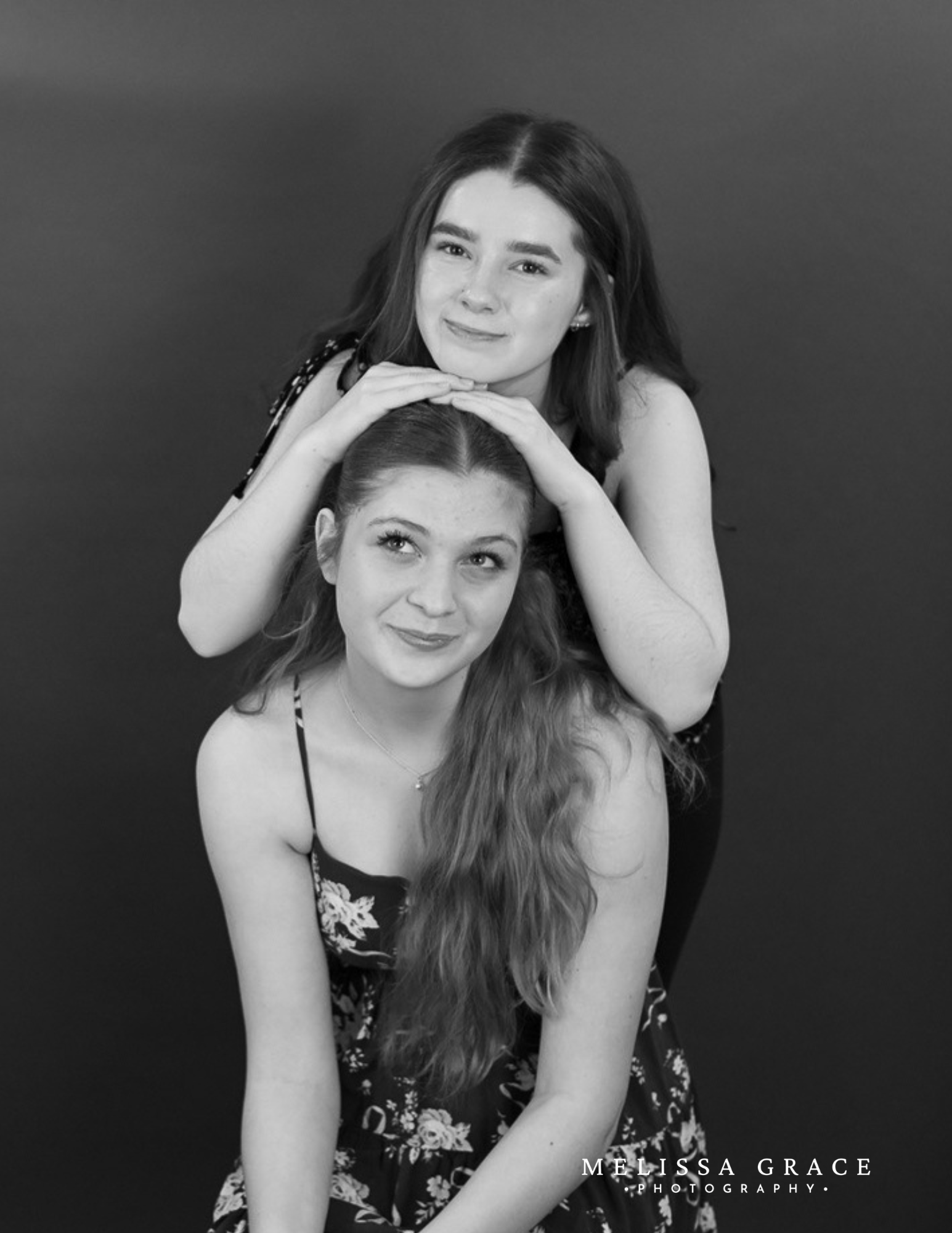 Black and white portrait of two young women, one with dark hair and the other with red hair, smiling and leaning on each other against a plain background. The photo includes text that reads 'Melissa Grace Photography' in the bottom right corner.