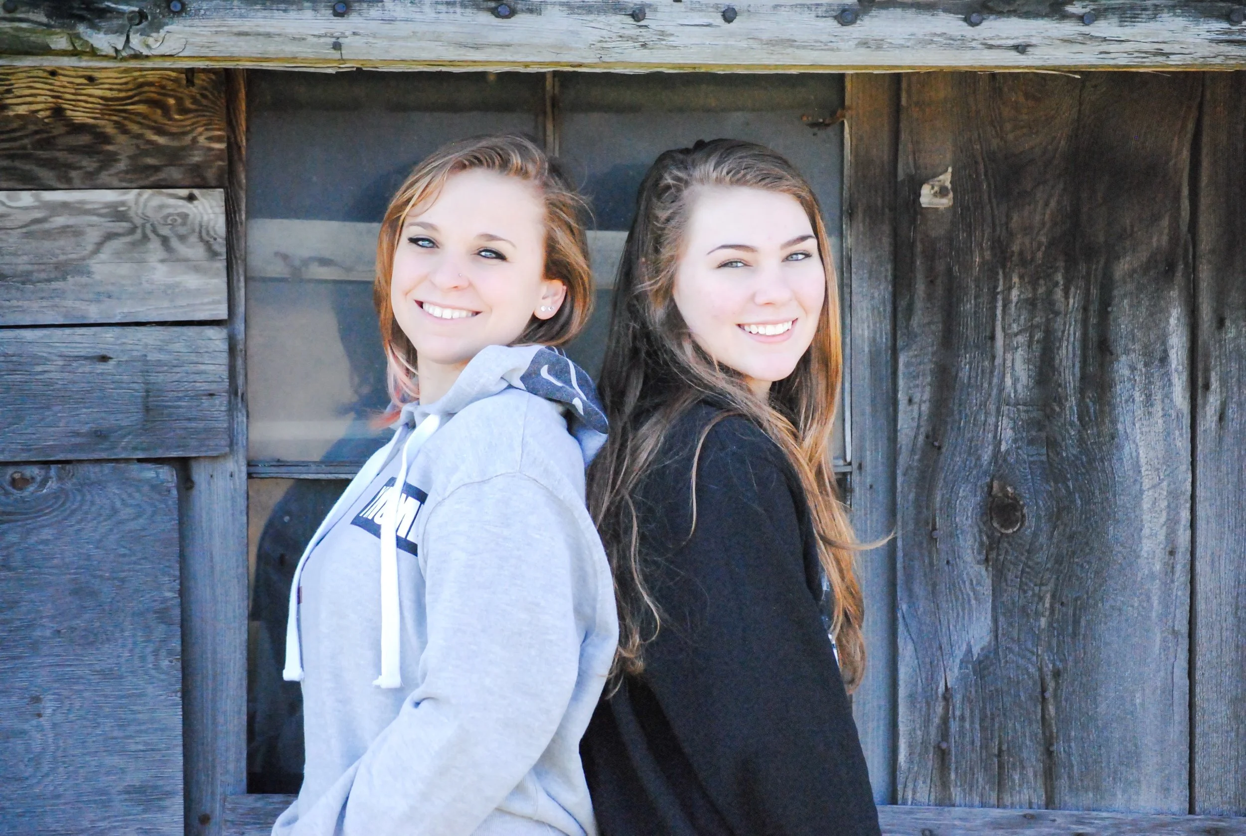 Two young women are standing back-to-back, smiling at the camera, against a rustic wooden wall.
