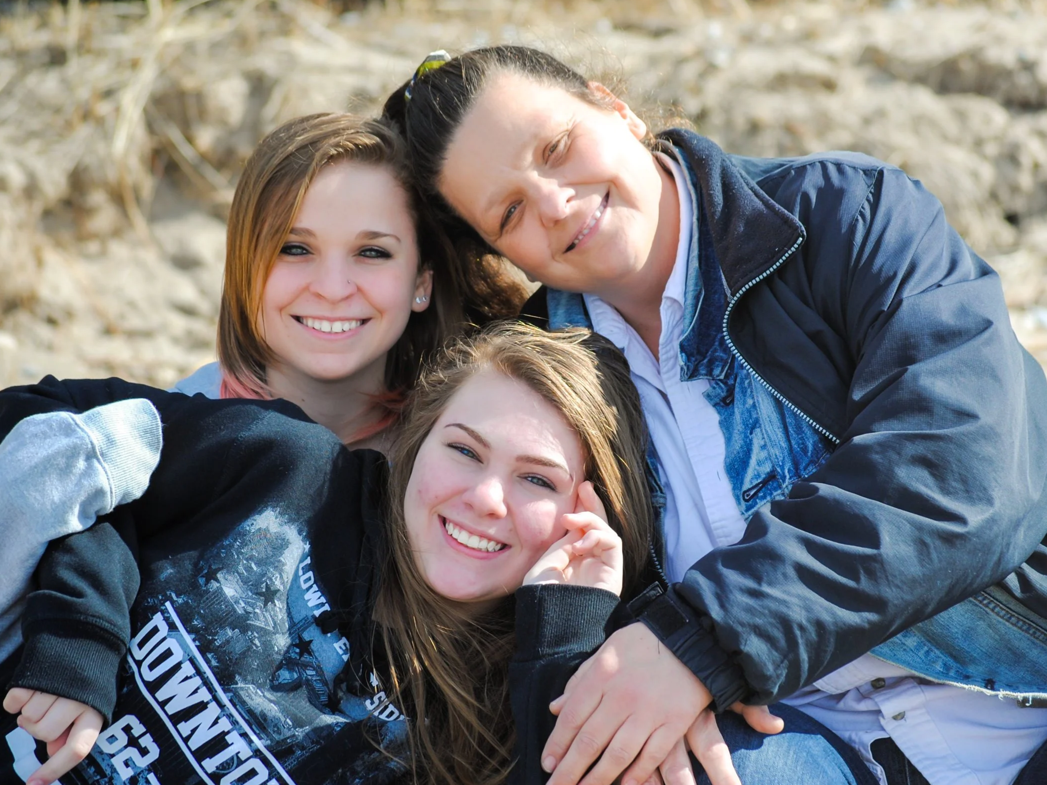 A group of smiling young women and an older woman taking a selfie outdoors with blurred natural background.