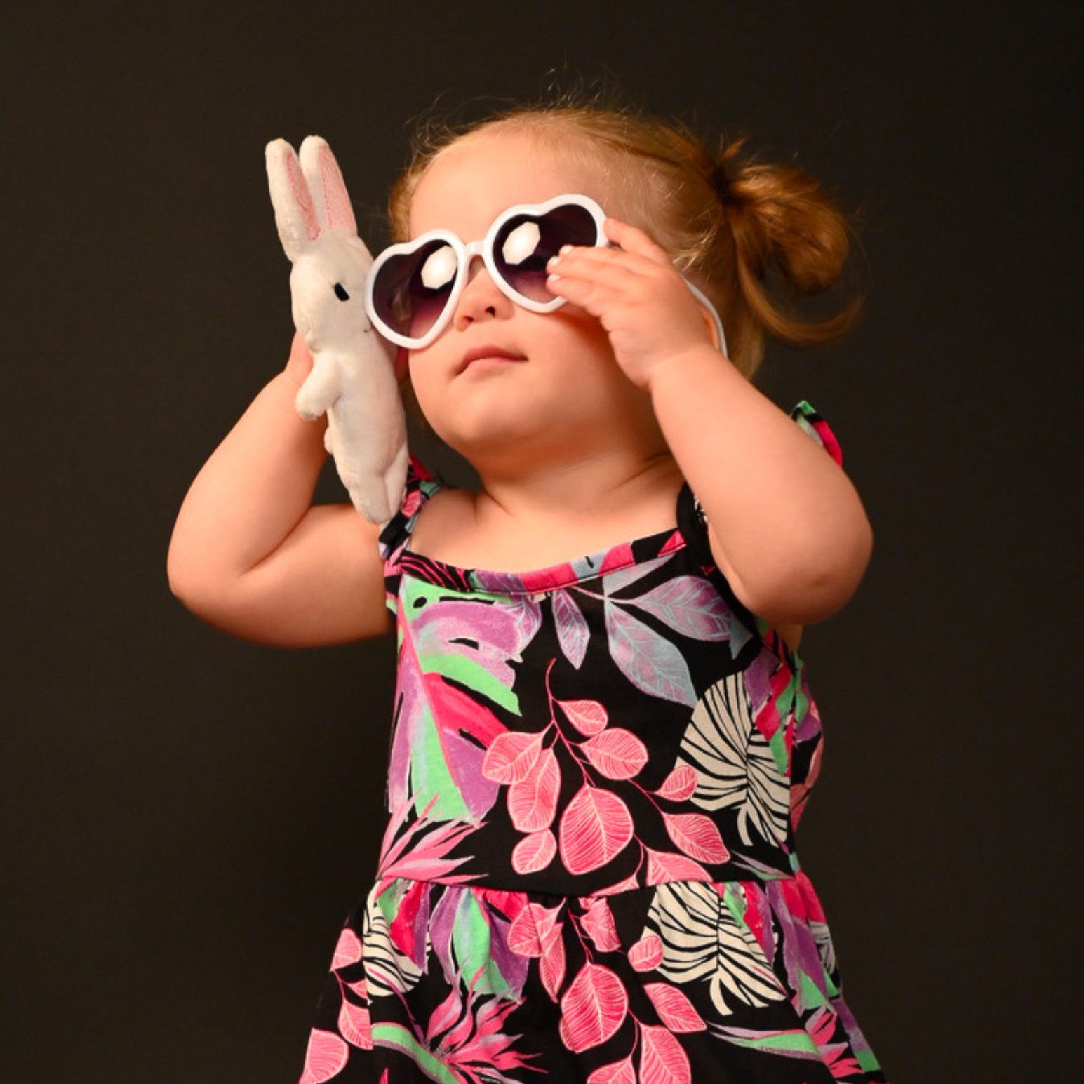 A young girl with red hair styled in buns, wearing large white sunglasses and a colorful dress with pink, green, and black leaf patterns. She is holding a white stuffed bunny toy and is against a dark background.