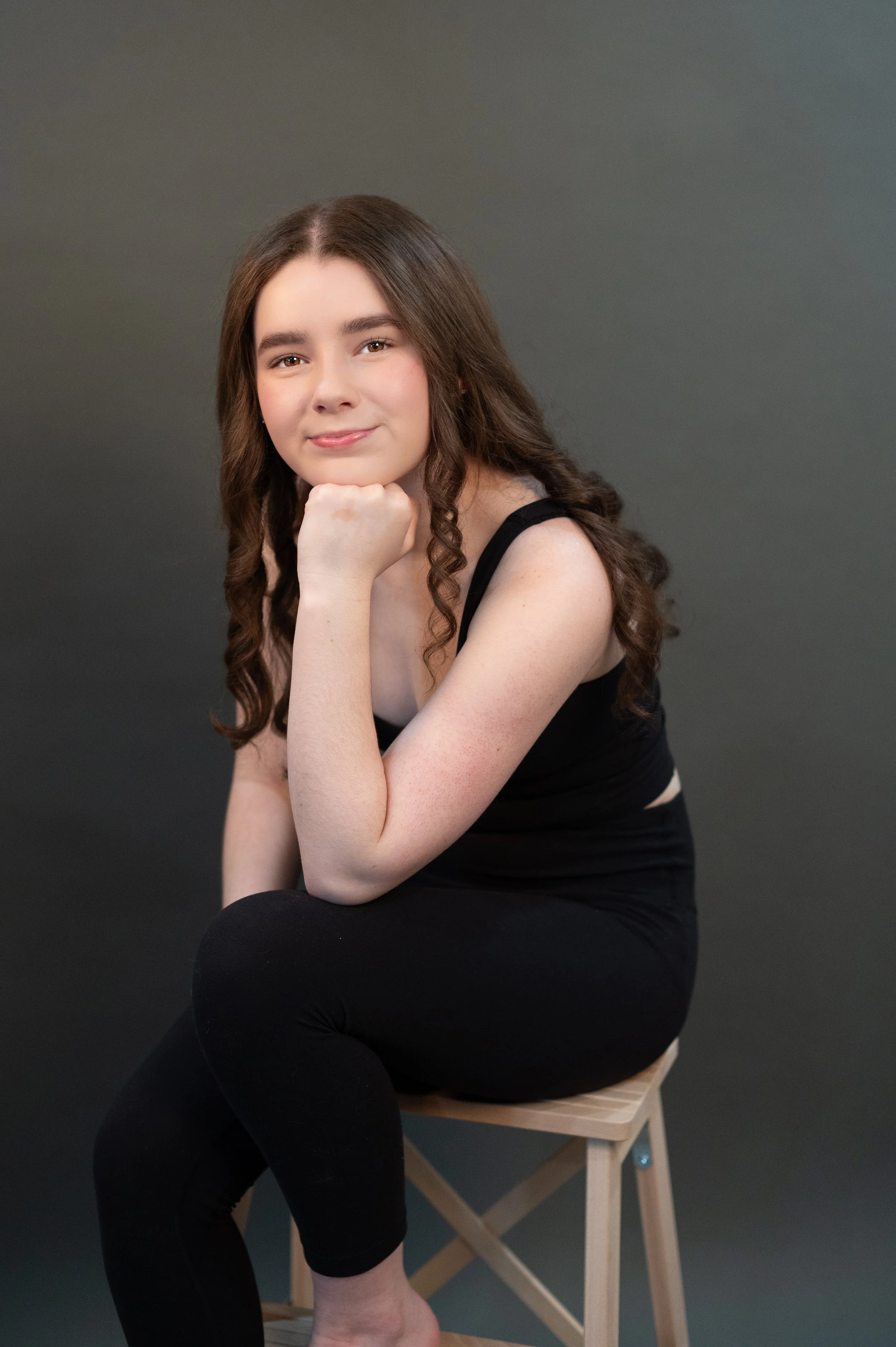 Young woman with long, wavy brown hair wearing a black sleeveless top, sitting on a wooden stool with one leg crossed over the other, resting her chin on her hand, against a plain gray background.