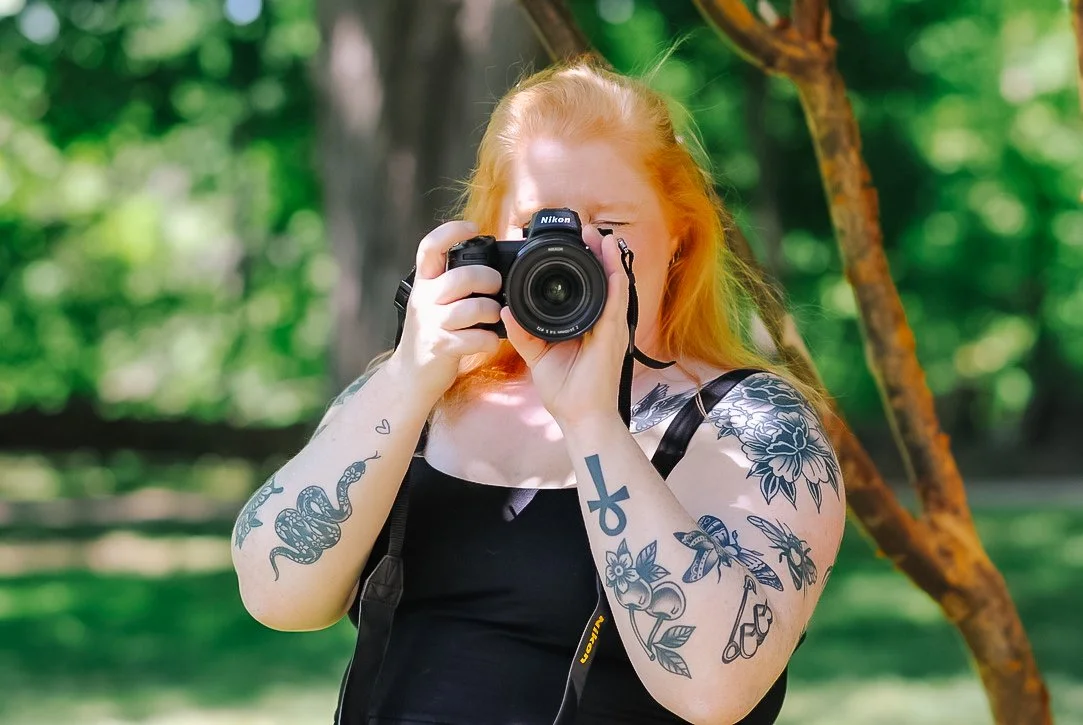A woman with long red hair taking a photo with a camera outdoors in a green park.